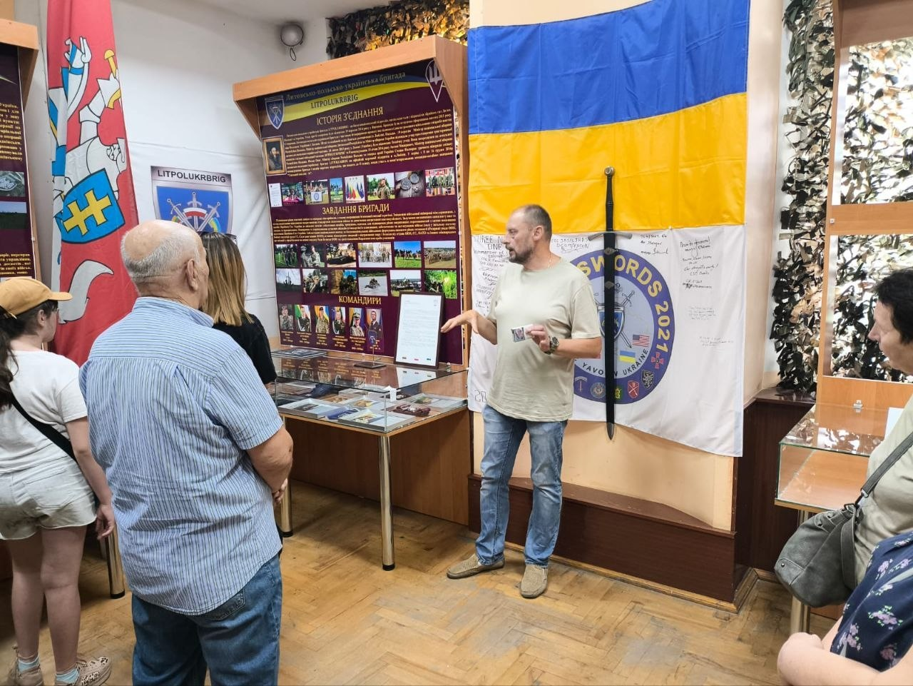 Екскурсовод розповідає переселенцям із Херсонщини про Литовсько-польсько-українську бригаду (ЛИТПОЛУКРБРИГ) у Будинку офіцерів у Житомирі на тлі інформаційного стенда, прапора бригади з мечем та прапора України з підписами.