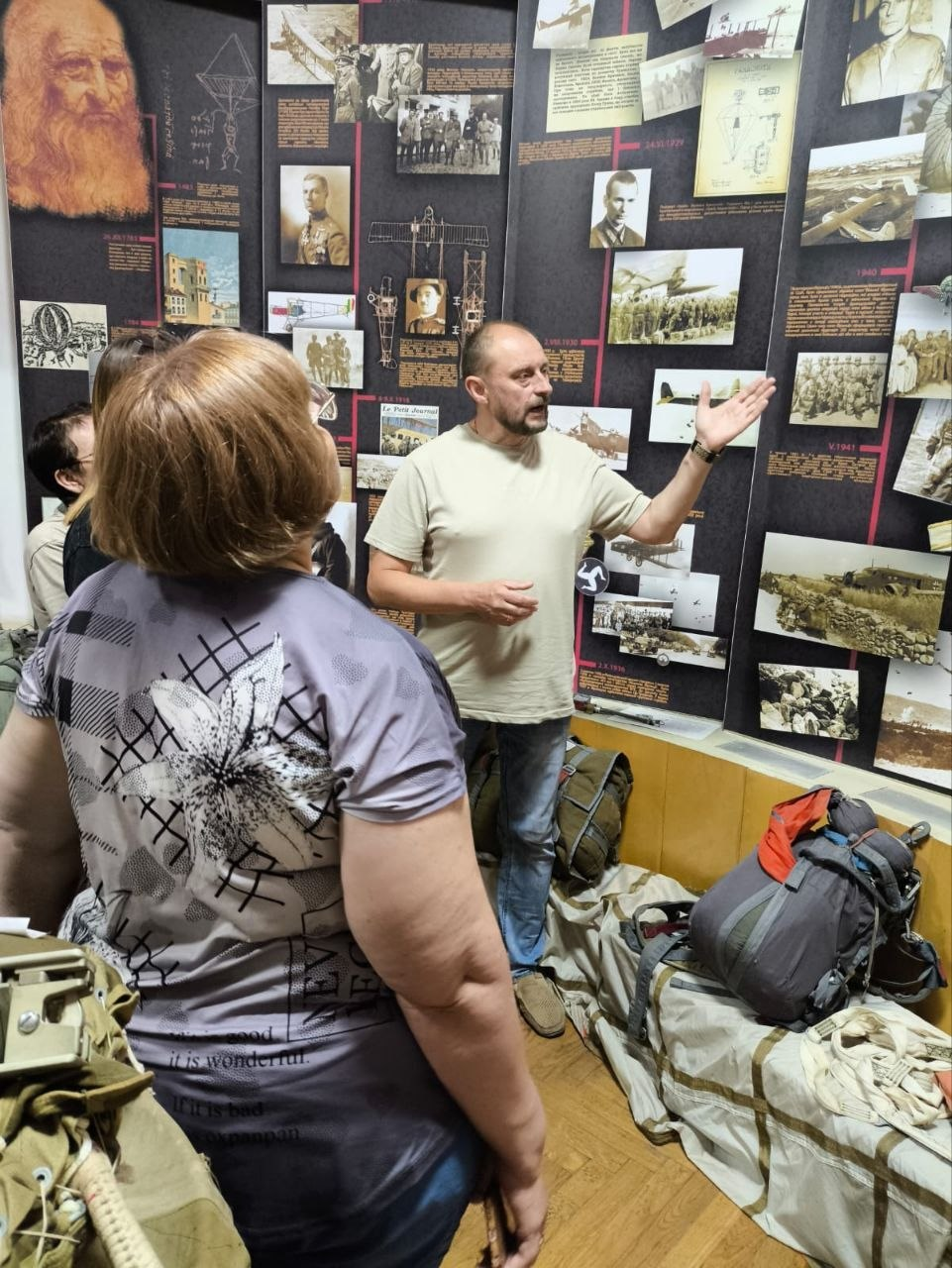 Екскурсовод у світлій футболці жестом показує на історичний стенд, розповідаючи переселенцям із Херсонщини про історію авіації та парашутного спорту в Житомирському Будинку офіцерів. Поруч на підлозі розкладено рюкзаки та елементи військового спорядження.