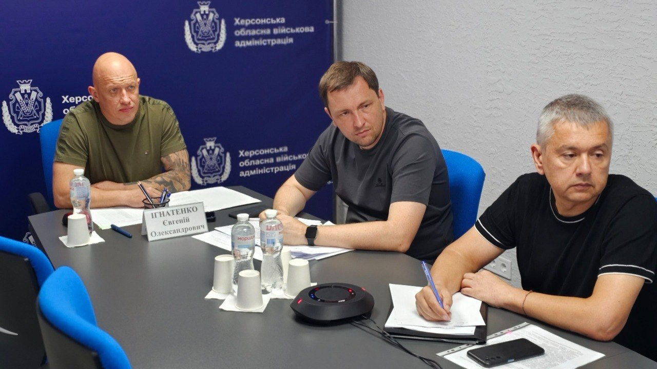 Заступник начальника Херсонської обласної військової адміністрації Євгеній Ігнатенко (ліворуч, у хакі-футболці), директор Департаменту з питань цивільного захисту та оборонної роботи Євген Пісоцький (у центрі, в сірій футболці) та посадовець у чорній футболці (праворуч) сидять за робочим столом під час засідання Координаційного штабу на тлі синього банера ХОВА.