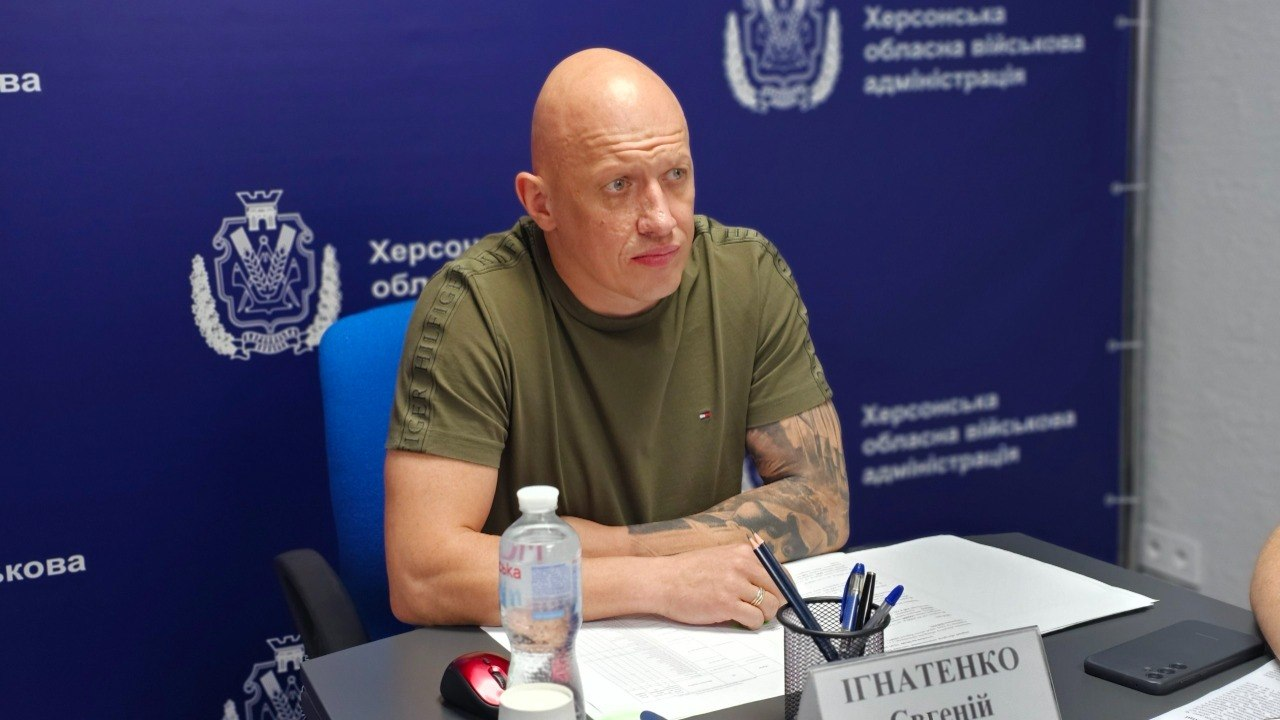 Заступник начальника Херсонської обласної військової адміністрації Євгеній Ігнатенко у футболці кольору хакі з татуюванням на лівій руці сидить за робочим столом під час засідання Координаційного штабу на тлі синього брендованого банера ХОВА.