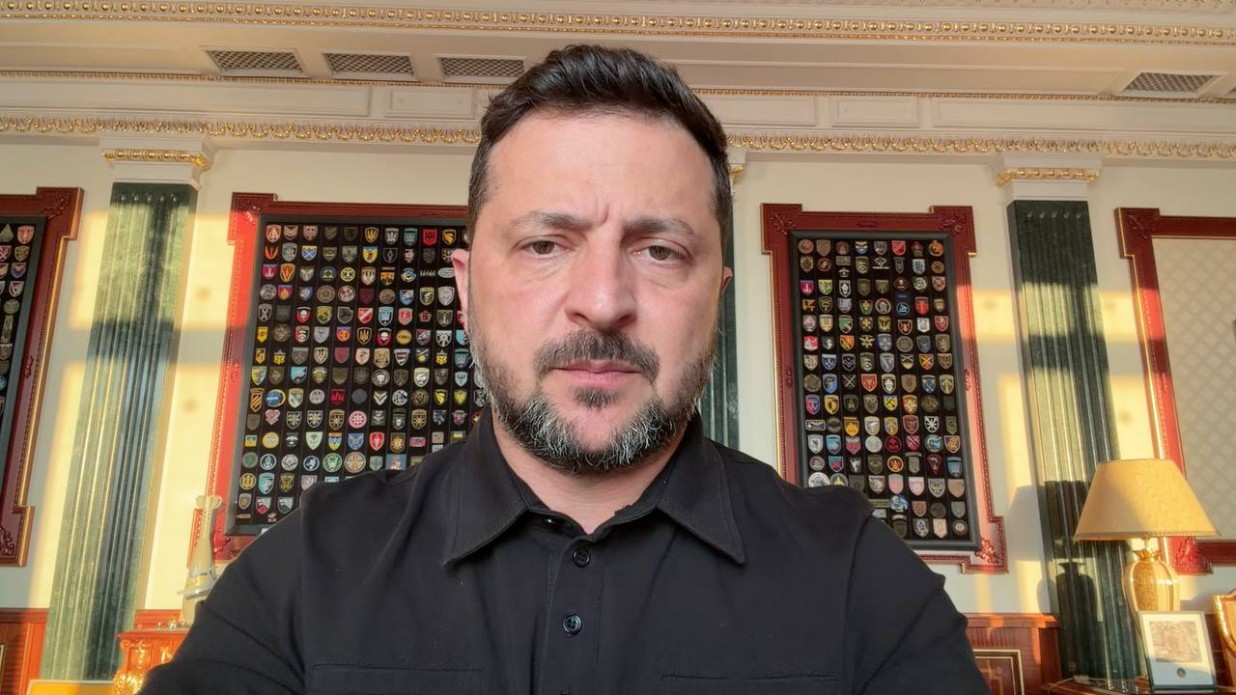 Президент України Володимир Зеленський у чорній сорочці звертається на камеру під час відеозвернення, сидячи у кабінеті на тлі стін із колонами та великих рамок із військовими шевронами.
