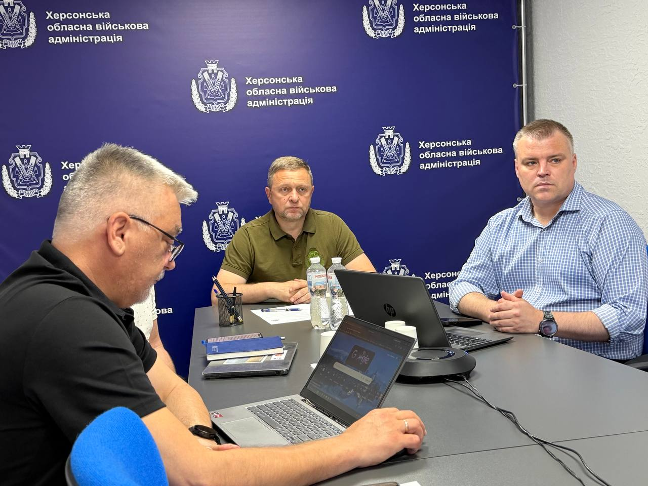 Три чоловіки беруть участь у робочій зустрічі за столом на тлі синього банера «Херсонська обласна військова адміністрація». По центру сидить перший заступник начальника Херсонської ОВА Дмитро Бутрій, праворуч від нього — директор Департаменту розвитку економіки ХОДА Артем Мішин, а на передньому плані ліворуч — чоловік, який працює за ноутбуком.