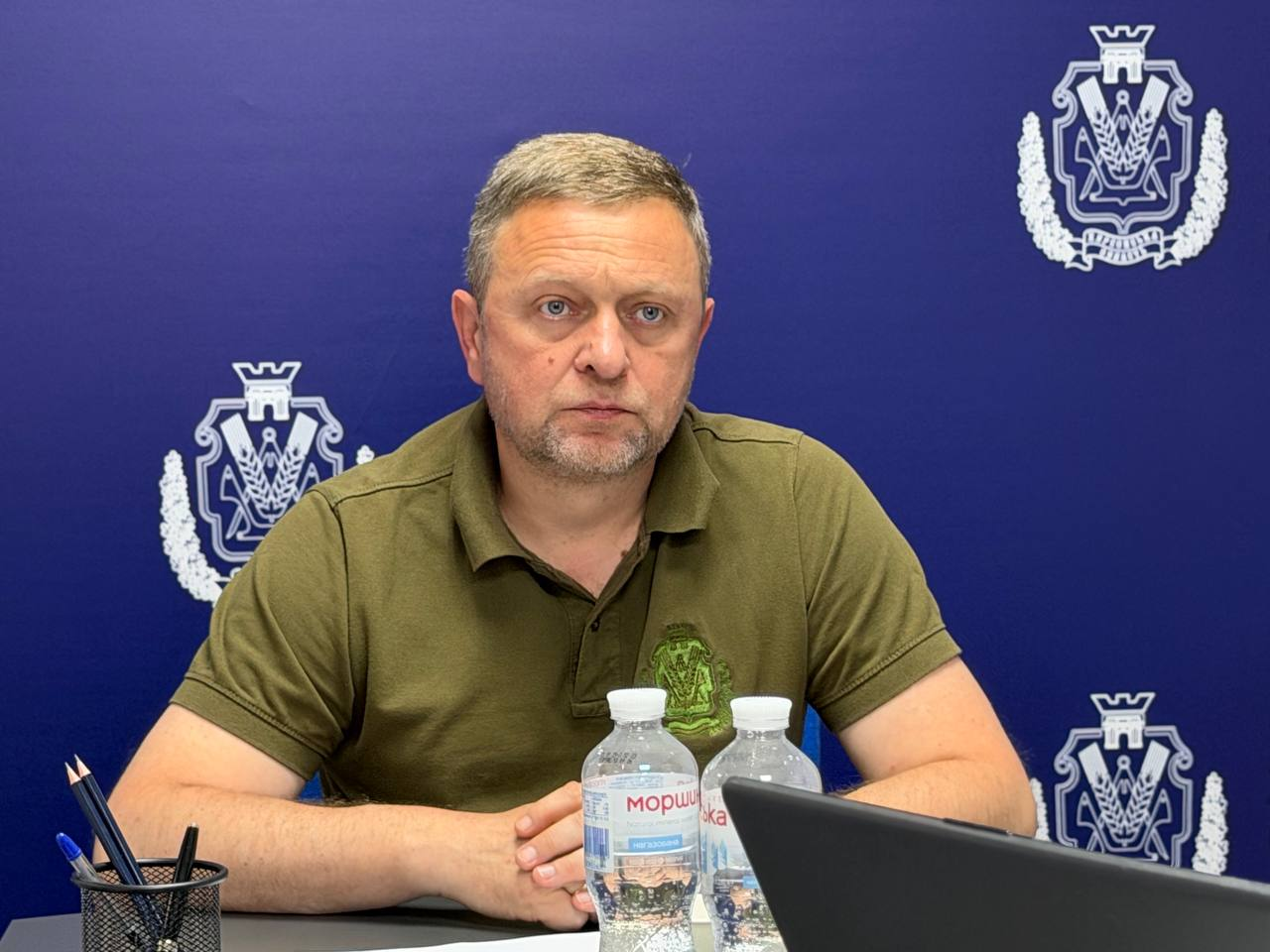 Перший заступник начальника Херсонської ОВА Дмитро Бутрій у футболці поло кольору хакі сидить за столом на тлі синього банера з гербами області.