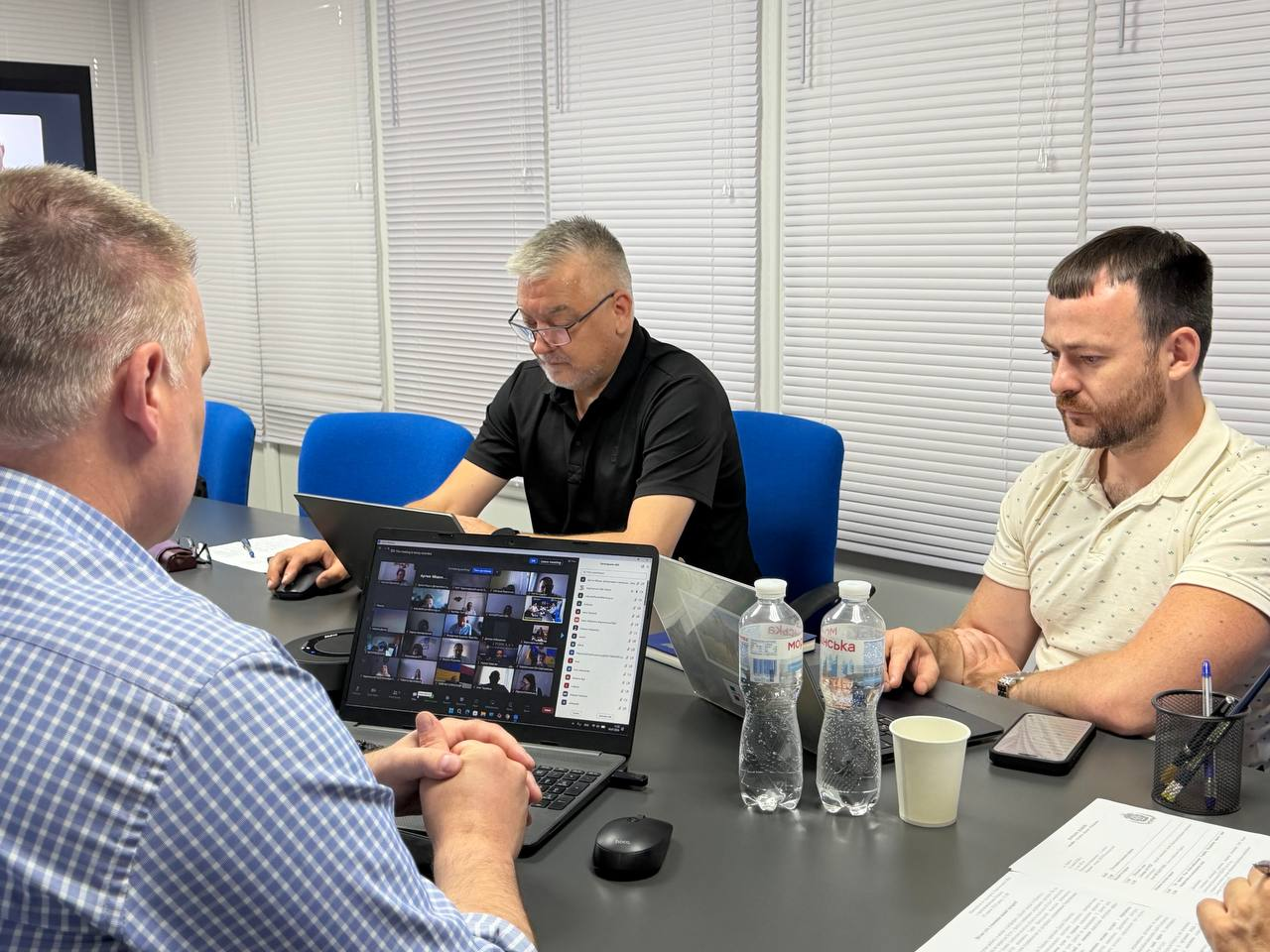 Три чоловіки беруть участь у робочій зустрічі за столом у кабінеті з жалюзі на вікнах. Ліворуч спиною до камери сидить директор Департаменту розвитку економіки ХОДА Артем Мішин, по центру — чоловік у чорній сорочці поло, а праворуч — керівник Агенції регіонального розвитку «Офіс євроінтеграції» Херсонської області Олег Кулакевич.