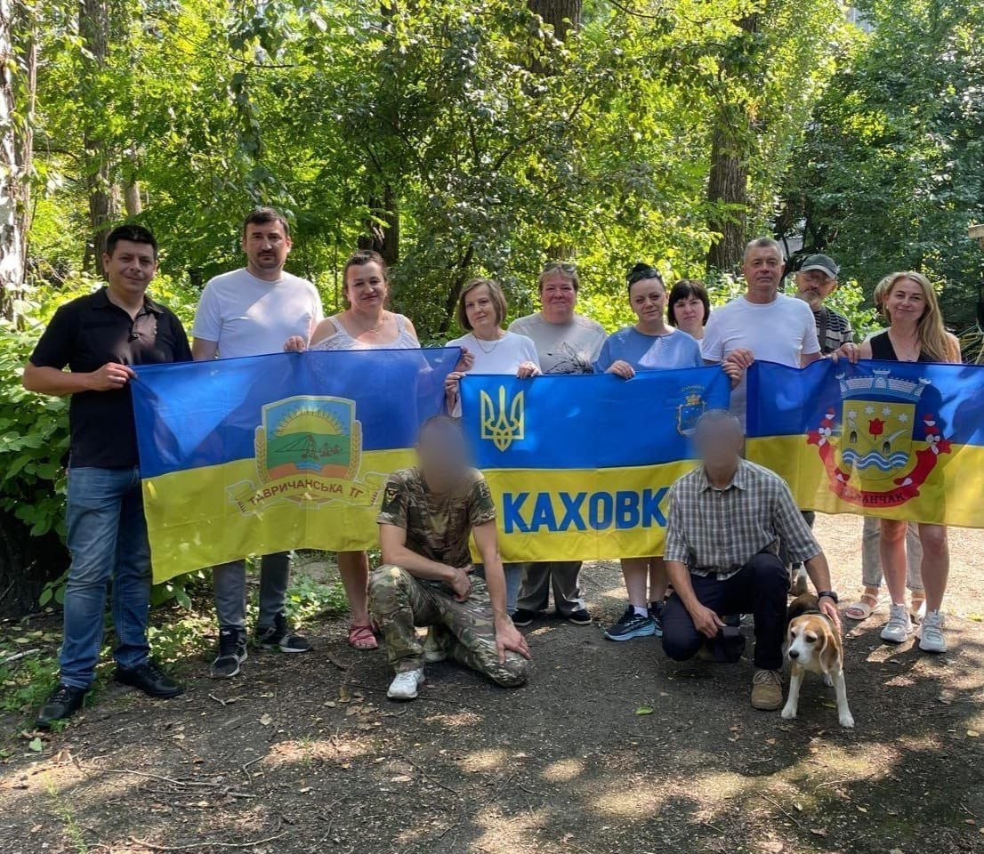 Команда координаційного центру «Вільні разом» на практичному базовому курсі від Київського обласного центру підготовки населення до національного спротиву. Група учасників стоїть на тлі дерев і тримає прапори Тавричанської ТГ, міста Каховка та Каланчака. Попереду присіли двоє чоловіків (обличчя розмиті), поруч із якими стоїть собака породи бігль.