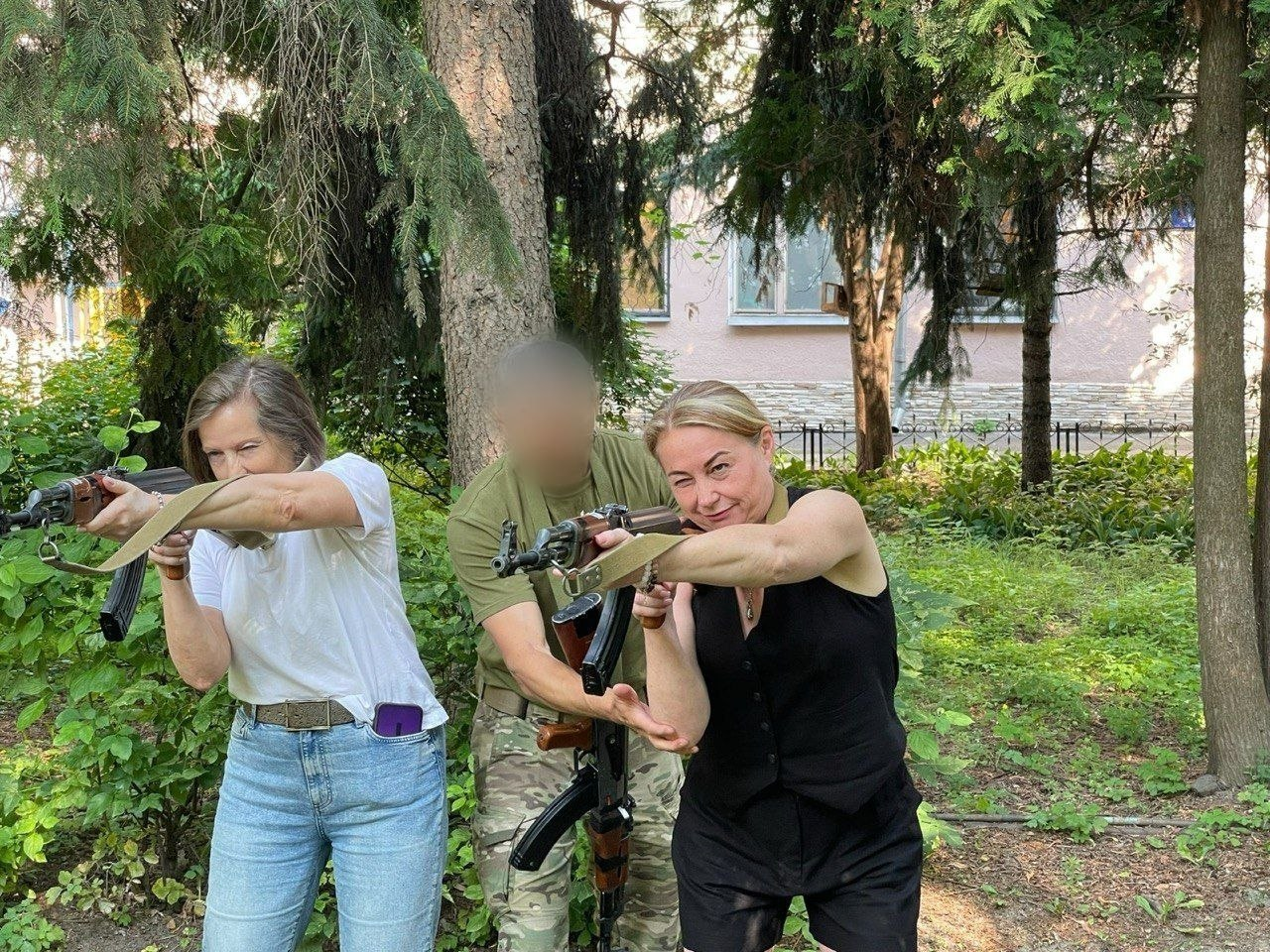 Дві жінки з команди координаційного центру «Вільні разом» на практичному базовому курсі відпрацьовують стійку та прицілювання з автоматами Калашникова на тлі дерев. По центру за ними стоїть інструктор у камуфляжній формі (обличчя розмите), який допомагає правильно тримати зброю.