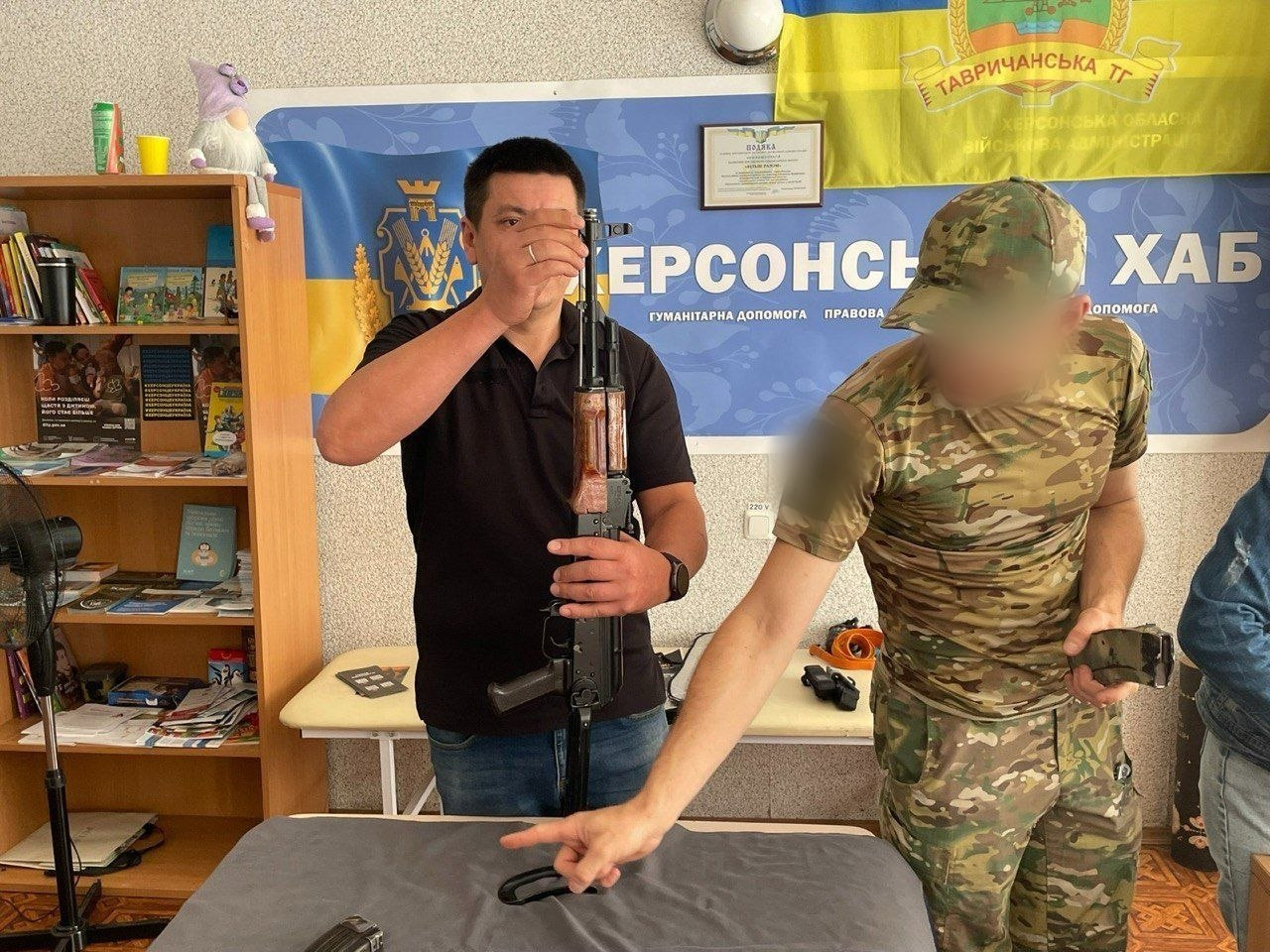 Учасник команди координаційного центру «Вільні разом» у чорній футболці відпрацьовує навички поводження з автоматом Калашникова під керівництвом інструктора у камуфляжі (обличчя розмите). Заняття проходить у приміщенні хабу на тлі синьо-жовтих банерів та прапора Тавричанської ТГ.