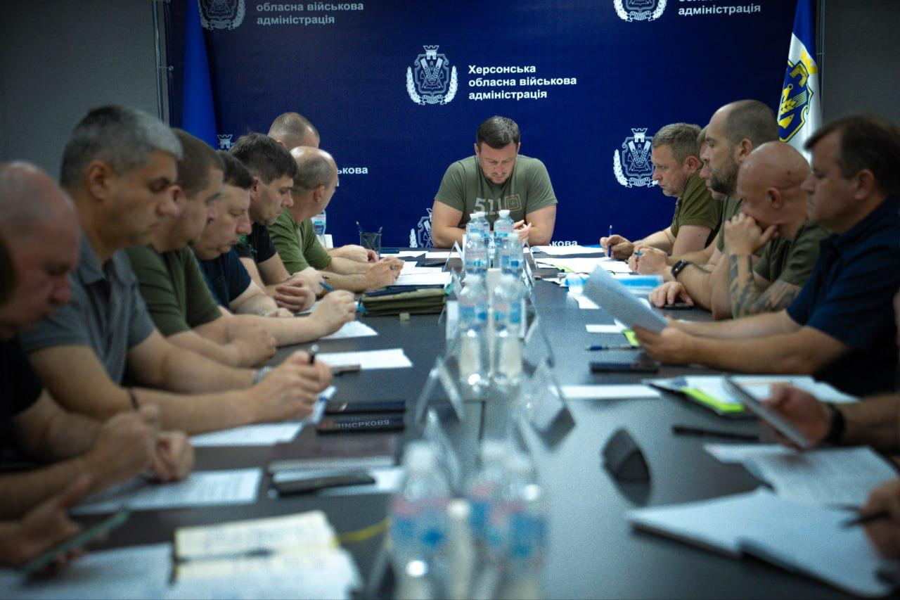 Начальник Херсонської ОВА Олександр Прокудін проводить засідання Ради оборони області за довгим столом на тлі синього брендованого банера. Праворуч від нього сидять його заступники: Дмитро Бутрій, Антон Самойленко, Олександр Толоконніков, Євгеній Ігнатенко та Володимир Клюцевський, а ліворуч та вздовж столу — інші посадовці, військові й представники силових структур.