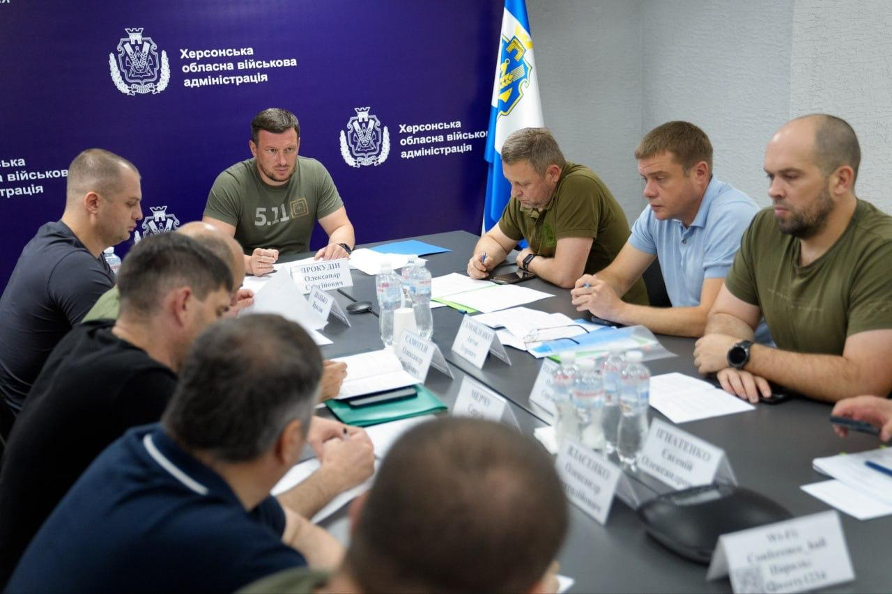 Начальник Херсонської ОВА Олександр Прокудін проводить засідання Ради оборони області. Перший ліворуч за столом сидить начальник Херсонської МВА Ярослав Шанько, а праворуч від очільника ОВА — його заступники Дмитро Бутрій, Антон Самойленко та Олександр Толоконніков. На столі перед учасниками наради розкладені папери та встановлені іменні таблички.