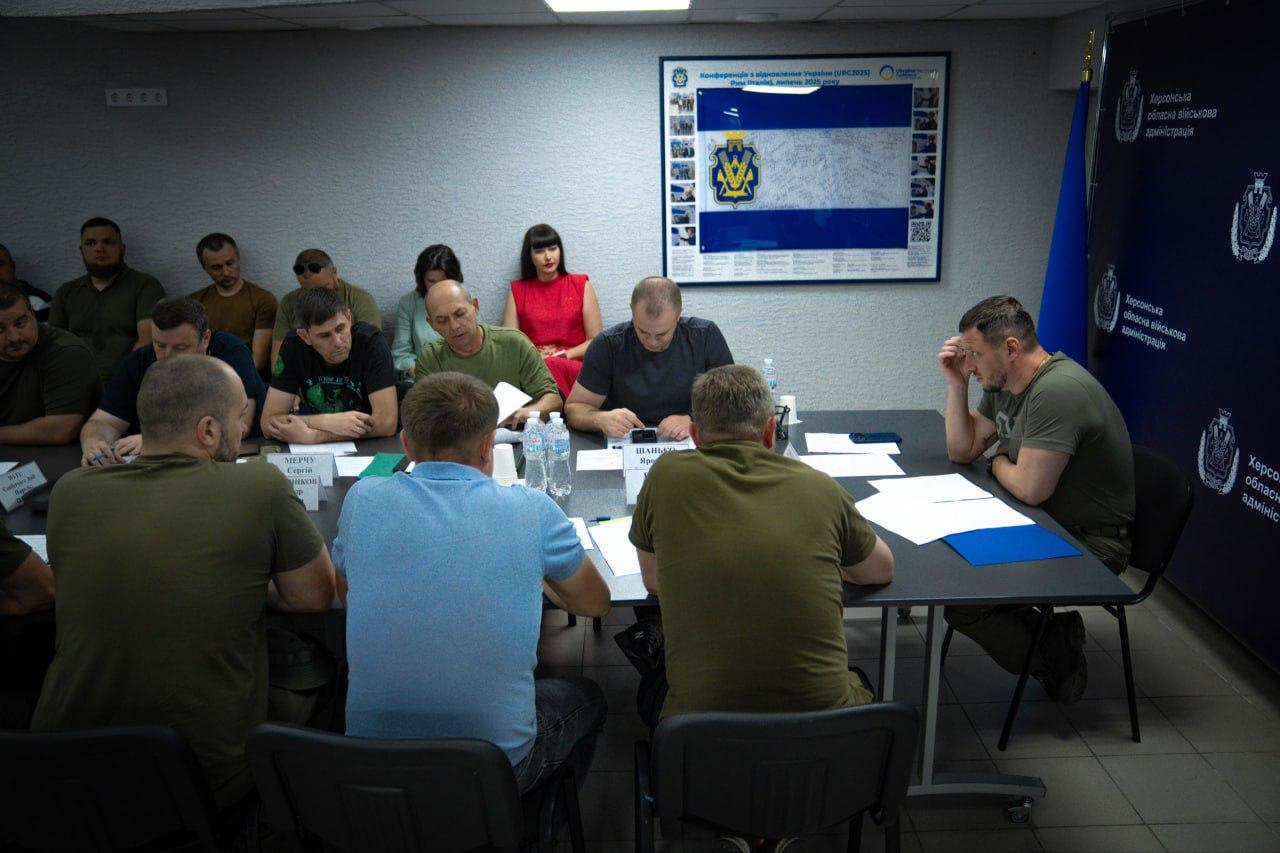 Начальник Херсонської ОВА Олександр Прокудін проводить засідання Ради оборони області за великим столом. На передньому плані (спиною до камери) сидять заступники очільника ОВА, серед яких Дмитро Бутрій, Антон Самойленко та Олександр Толоконніков. Навпроти Дмитра Бутрія обличчям до камери сидить начальник Херсонської МВА Ярослав Шанько у темно-сірій футболці, а навколо за столом та вздовж стін присутні інші посадовці й представники силових структур.