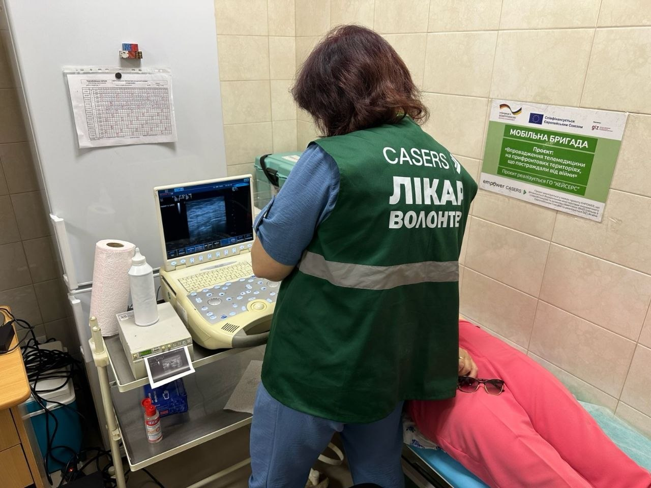 Лікарка мобільної бригади ГО «CASERS» у зеленому жилеті з написом «ЛІКАР ВОЛОНТЕР» спиною до камери проводить ультразвукове дослідження (УЗД) пацієнту, який лежить на кушетці. Вона налаштовує портативний апарат УЗД, а на стіні праворуч висить плакат проєкту «Мобільна бригада».