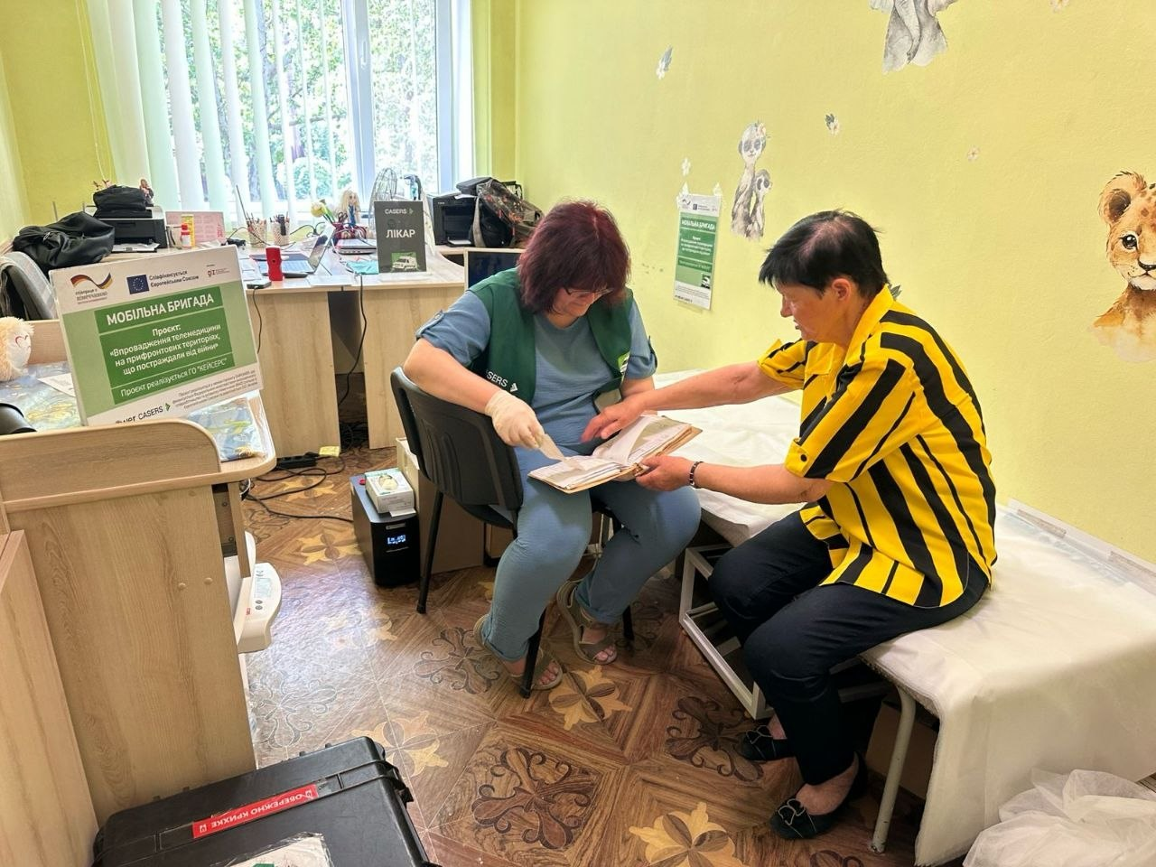 Лікарка мобільної бригади ГО «CASERS» у зеленому жилеті сидить поруч із пацієнткою в жовто-чорній сорочці та разом із нею переглядає медичну картку. Консультація проходить у світлому кабінеті, а на передньому плані ліворуч на столі стоїть інформаційний плакат проєкту «Мобільна бригада».