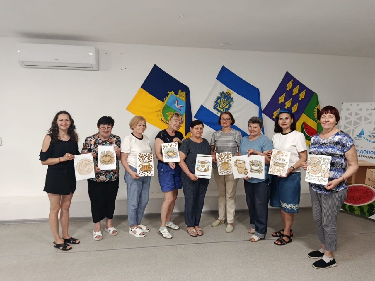 Переселенки з Херсонщини на ароматерапевтичному заході у координаційному центрі «Вільні разом» у Черкасах. Дев'ять жінок стоять у приміщенні та тримають у руках свої малюнки, створені кавою. Захід організовано у співпраці з Чаплинською та Долматівською СВА, а на стіні позаду учасниць висять прапори громад.