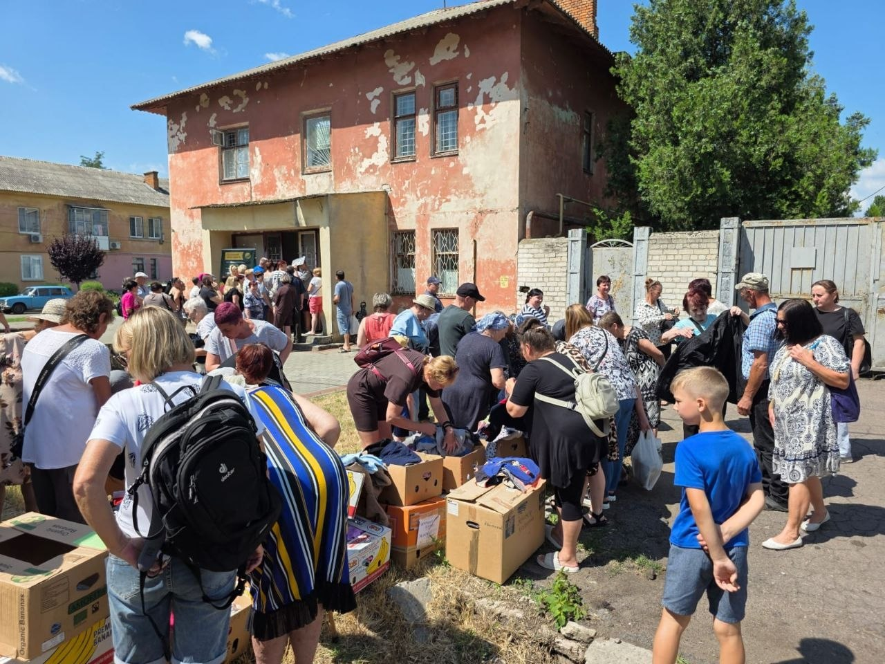 Велика група людей зібралася на подвір'ї біля двоповерхової будівлі з облупленою червоною фарбою під час виїзної видачі допомоги від координаційного центру «Вільні разом». На передньому плані люди перебирають одяг та речі, розкладені у великих картонних коробках, а на задньому плані видно чергу мешканців біля входу.