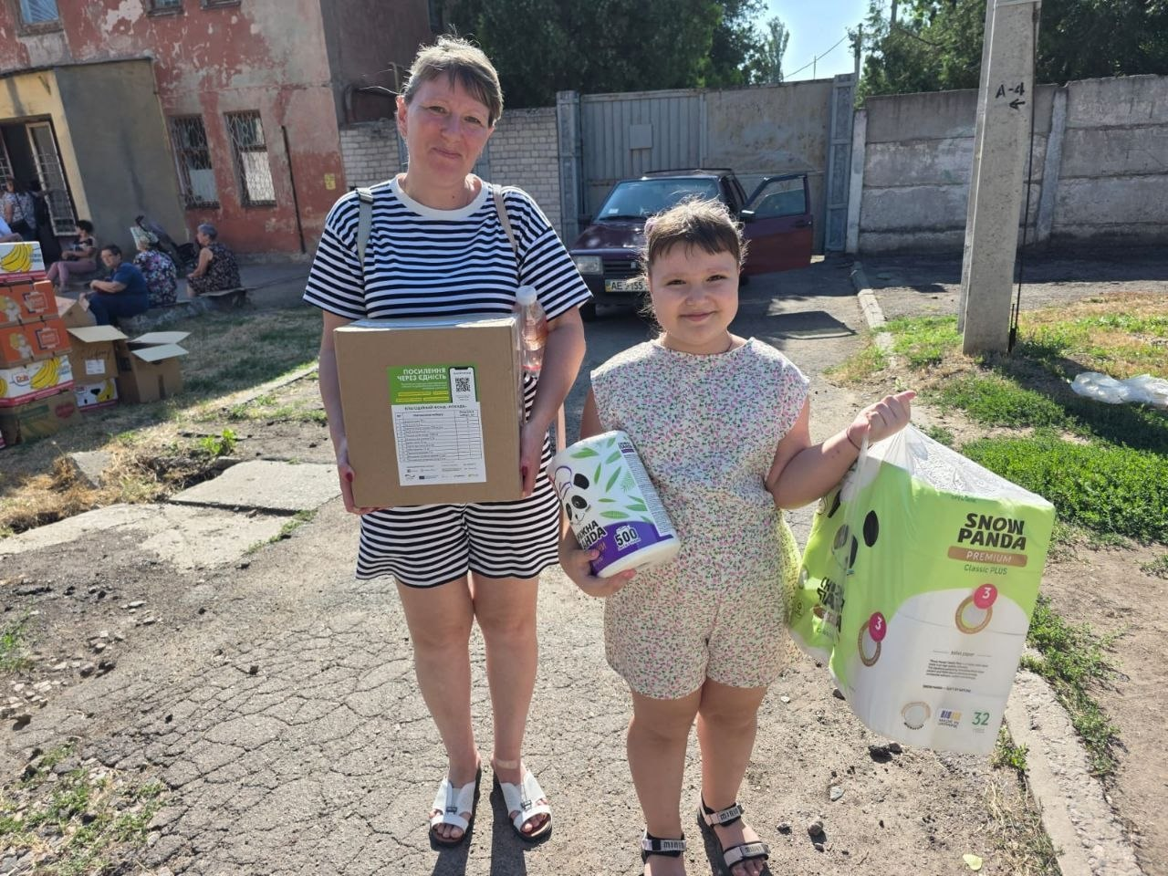 Жінка в смугастій сукні та дівчинка посміхаються, тримаючи в руках отриману гуманітарну допомогу на вулиці. Жінка тримає велику картонну коробку з наліпкою «Посилення через єдність», а дівчинка — велику упаковку туалетного паперу «Snow Panda» та рулон паперових рушників під час виїзної видачі від координаційного центру «Вільні разом».