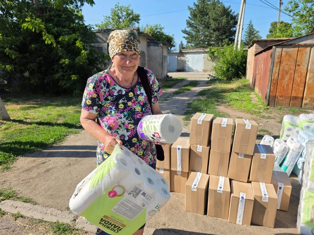 Літня жінка у квітчастій футболці та хустці посміхається, тримаючи в руках отриману гуманітарну допомогу на вулиці. Вона стоїть на тлі складених картонних коробок і упаковок гігієнічних засобів, тримаючи велику упаковку туалетного паперу «Сніжна Панда» та рулон паперових рушників під час виїзної видачі від координаційного центру «Вільні разом».