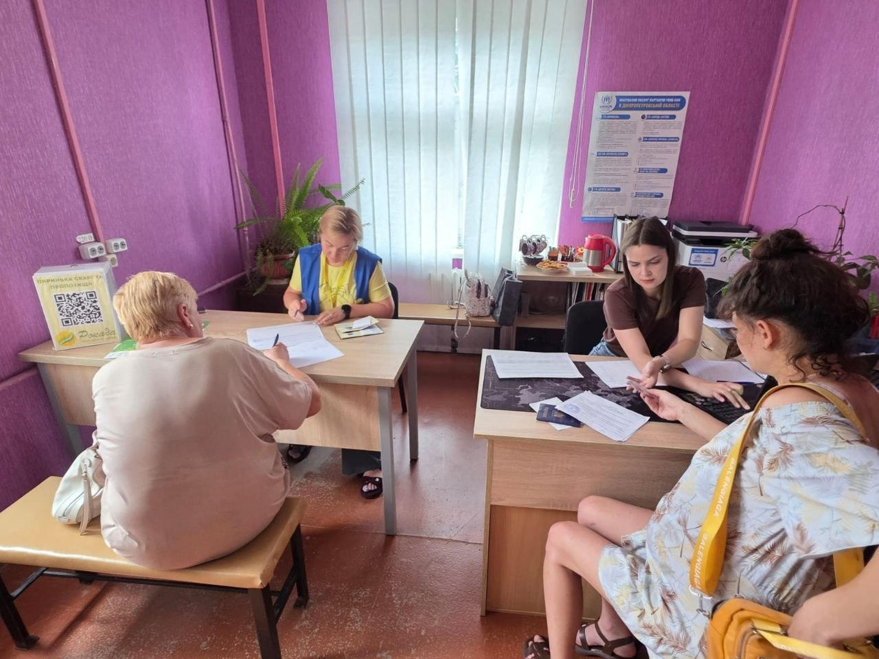 Дві працівниці координаційного центру «Вільні разом» у кабінеті з рожевими стінами приймають громадян і допомагають їм заповнювати документи. Ліворуч за столом жінка в синьо-жовтому жилеті працює з літньою відвідувачкою, а праворуч інша співробітниця консультує молоду жінку, на столі перед якими лежать паспорти та бланки.