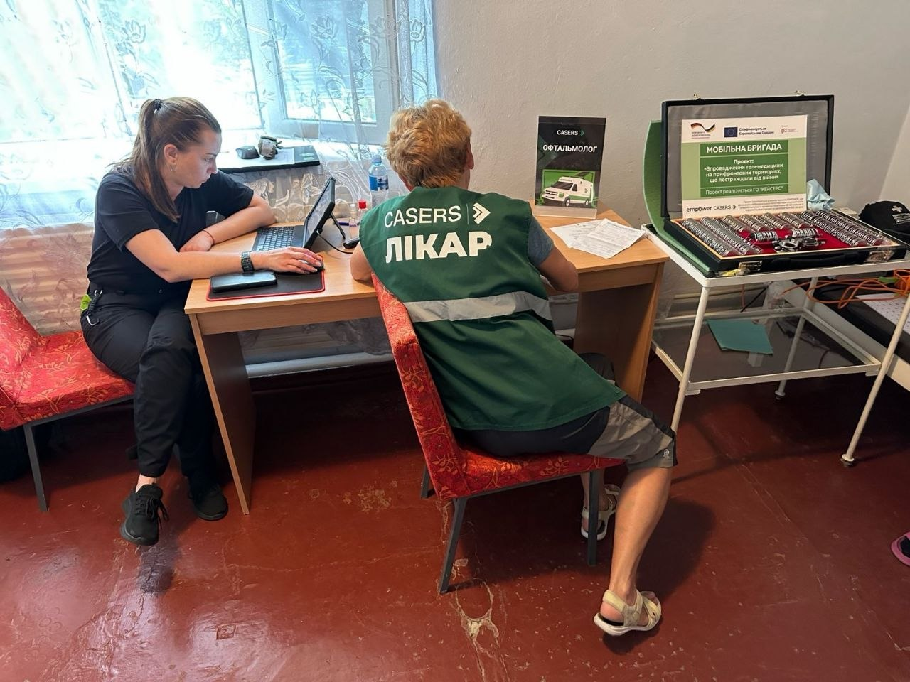 Два медичних працівники мобільної бригади ГО «CASERS» працюють у кабінеті. Одна жінка вносить дані у ноутбук, а інша, у фірмовому зеленому жилеті з написом «CASERS ЛІКАР», сидить поруч. На столі та підставці розміщено діагностичне обладнання, інформаційні матеріали та відкриту валізу з набором лінз офтальмолога.