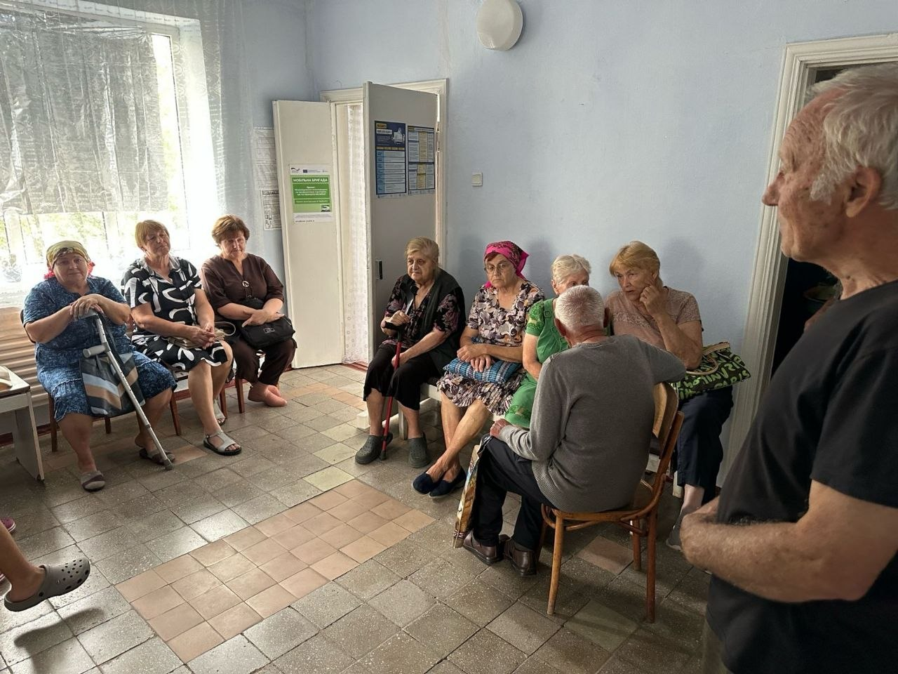 Місцеві мешканці старшого віку очікують у коридорі на прийом до спеціалістів мобільної бригади ГО «CASERS». Люди сидять на стільцях уздовж стін та спілкуються між собою, а на дверях кабінету позаду розміщено інформаційні плакати проєкту.