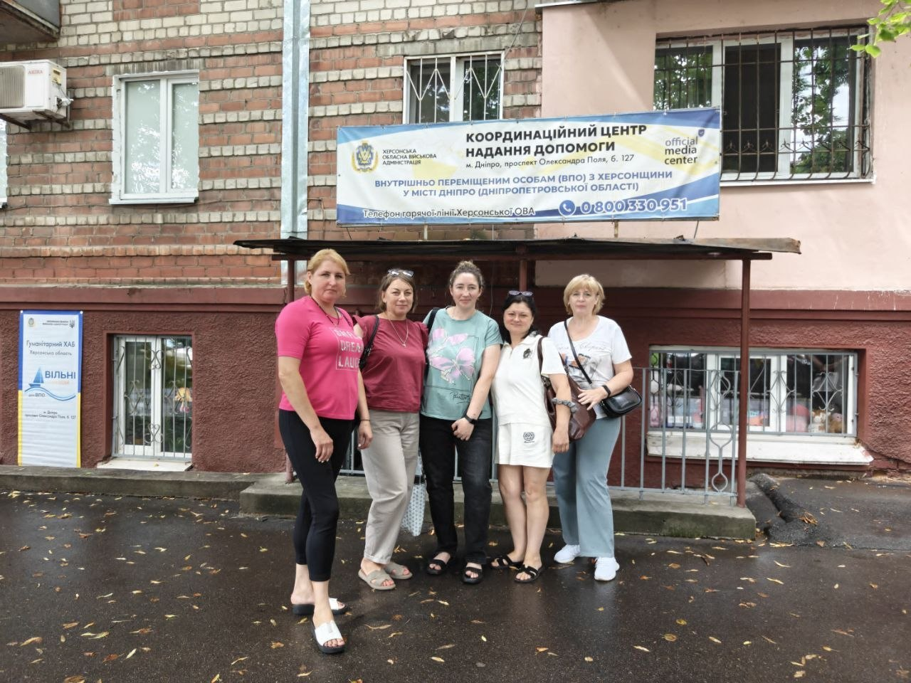 П'ять жінок стоять перед входом до Координаційного центру надання допомоги ВПО з Херсонщини у місті Дніпро. Над ґанком будівлі закріплено інформаційний банер із контактами гарячої лінії, а ліворуч від дверей розташовано стенд гуманітарного хабу «Вільні разом».