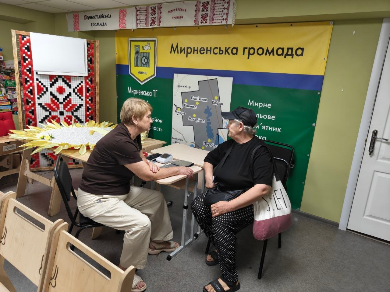 Медичний працівник проводить прийом пацієнтки старшого віку в приміщенні хабу Мирненської громади. Жінки сидять за столом одна навпроти одної, обговорюючи результати вимірювання тиску за допомогою тонометра, а на стіні позаду них розміщено великий інформаційний банер із мапою «Мирненська громада».