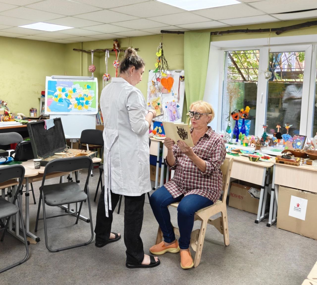 Лікар-офтальмолог проводить перевірку зору пацієнтки в облаштованому кабінеті хабу. Медичний працівник у білому халаті стоїть поруч із жінкою, яка сидить на стільці у спеціальних пробних окулярах для підбору лінз і читає текст із книги, а на задньому плані видно навчальні матеріали, дитячі вироби та мапу України.