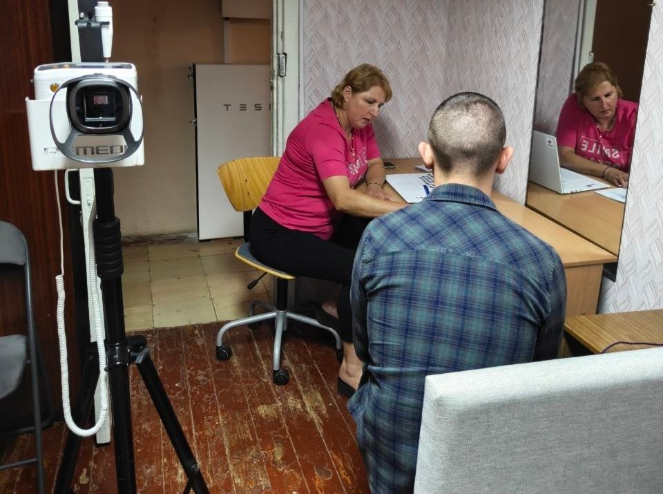 Лікар Нововоронцовської центральної районної лікарні проводить прийом пацієнта в координаційному центрі в Дніпрі. Жінка в рожевій футболці робить записи за робочим столом, спиною до неї на передньому плані сидить чоловік у карпованій сорочці, а ліворуч на штативі встановлено мобільне медичне діагностичне обладнання.