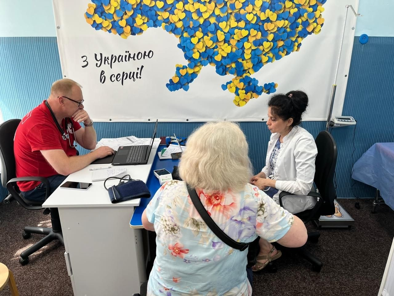 Медичне обстеження та консультація літньої пацієнтки зі світлим волоссям у квітковій футболці фахівцями мобільної бригади Червоного Хреста України на тлі плаката з картою України та написом «З Україною в серці!».