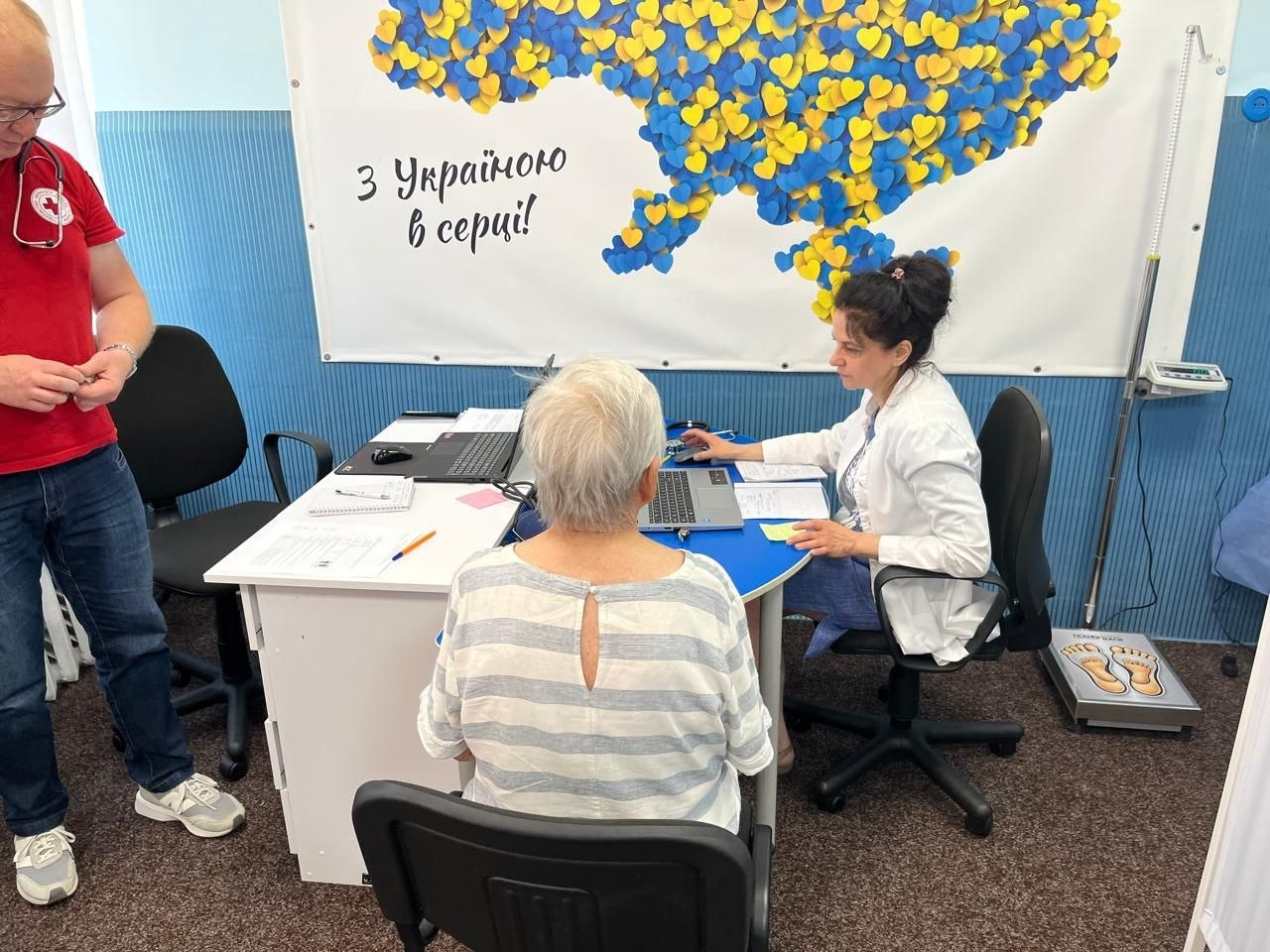 Медична консультація літньої пацієнтки у світлій смугастій кофті фахівцями мобільної бригади Червоного Хреста України. Жінка-медик у білому халаті сидить за столом із ноутбуком, а ліворуч стоїть лікар у червоній футболці з логотипом організації. На стіні розміщено банер із картою України та написом «З Україною в серці!».
