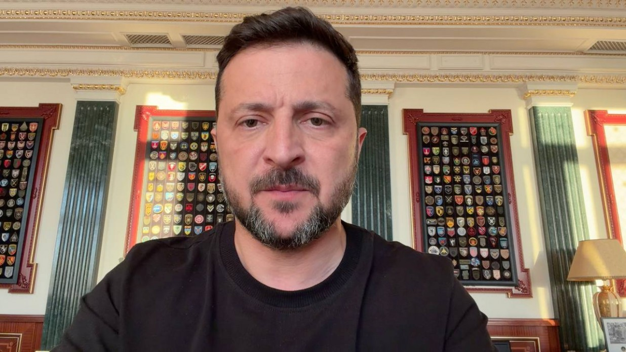 Президент України Володимир Зеленський під час відеозвернення. Він одягнений у чорний світшот та зафіксований на тлі кабінету з колонами, де на стінах розміщені стенди з великою кількістю шевронів українських військових підрозділів.