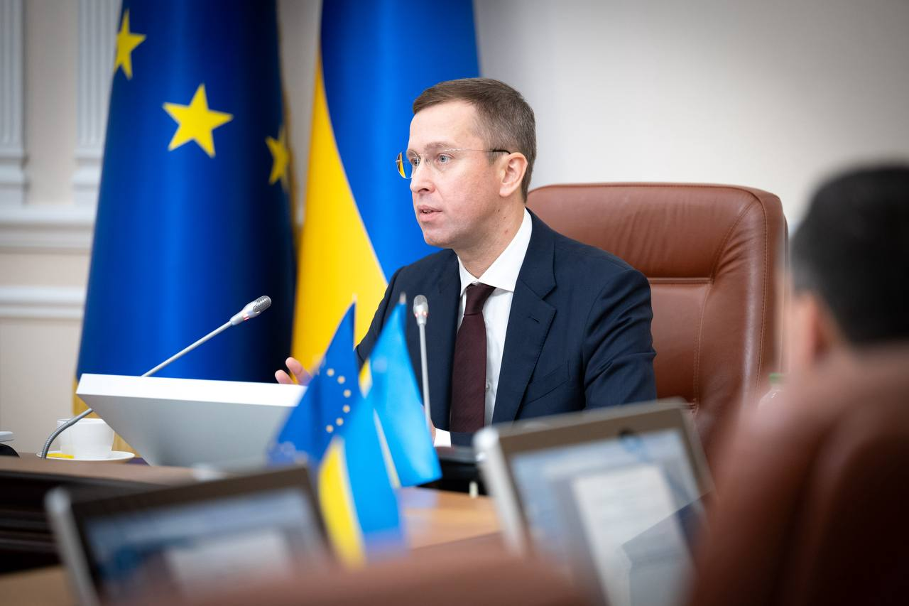 Прем'єр-міністр України Сергій Корецький виступає під час засідання Кабінету Міністрів. Глава Уряду одягнений у темно-синій костюм, білу сорочку та краватку, сидить у шкіряному кріслі за столом із мікрофонами на тлі державних прапорів України та Європейського Союзу.