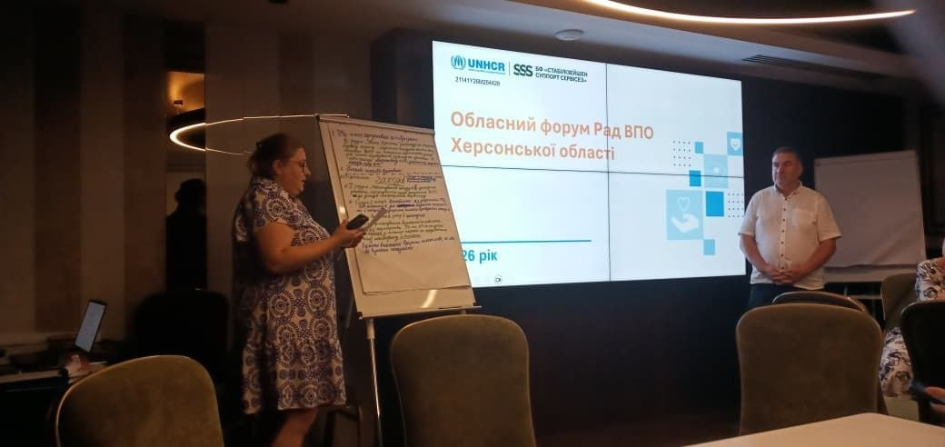 Презентація на Обласному форумі Рад ВПО Херсонської області. Жінка-спікер стоїть біля фліпчарта з рукописними нотатками та дивиться в телефон, а чоловік стоїть поруч біля великого екрана з офіційним слайдом заходу, логотипами UNHCR та БФ «Стабілізейшен Суппорт Сервісез» і написом «2026 рік».