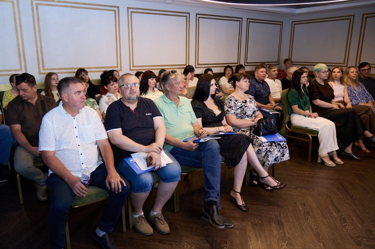 Учасники Обласного форуму Рад ВПО Херсонської області сидять у конференц-залі та уважно слухають виступ. Люди різного віку розмістилися на стільцях у кілька рядів, тримаючи в руках папки з робочими матеріалами та блокноти синього кольору.