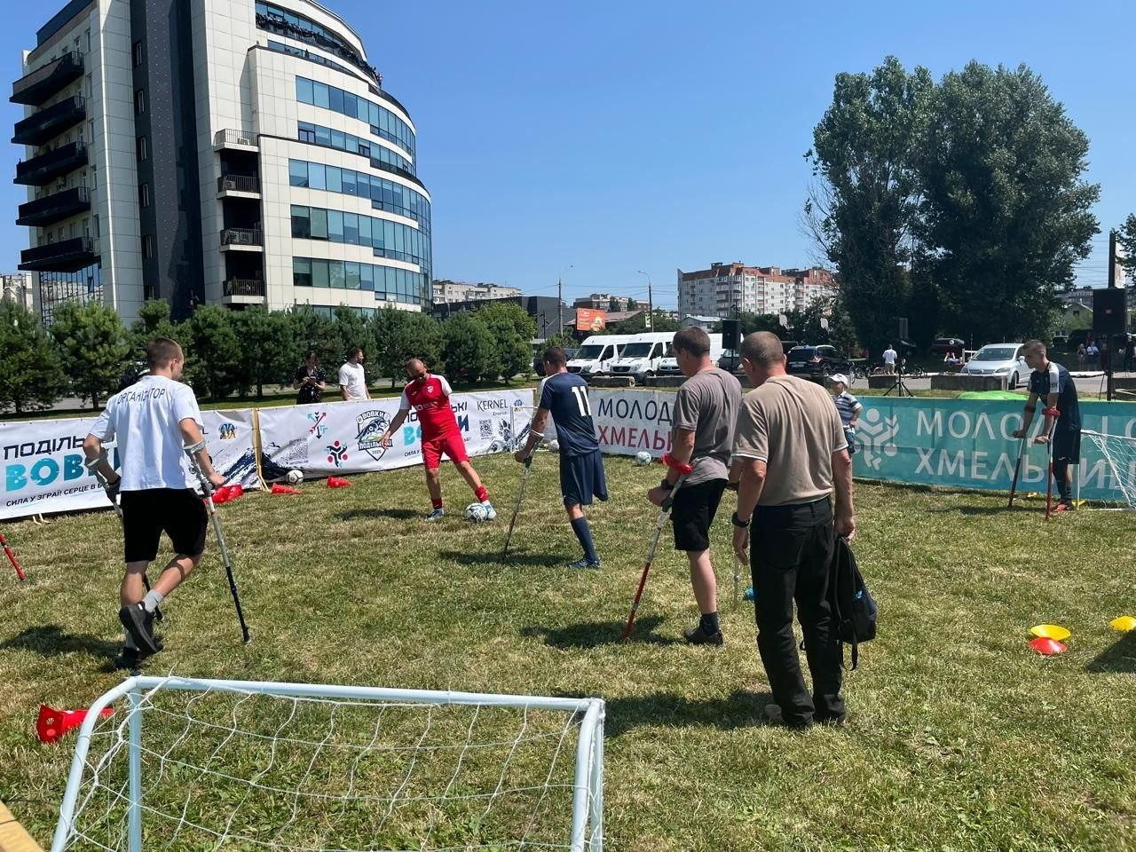 Зона адаптивного футболу на фестивалі «Ветеран Сіті Спорт 2026» у Хмельницькому. Гравці на милицях тренуються та передають м'яч на зеленому газоні Молодіжного парку. На тлі встановлені банери заходу з логотипами, а також видно сучасну багатоповерхову будівлю.