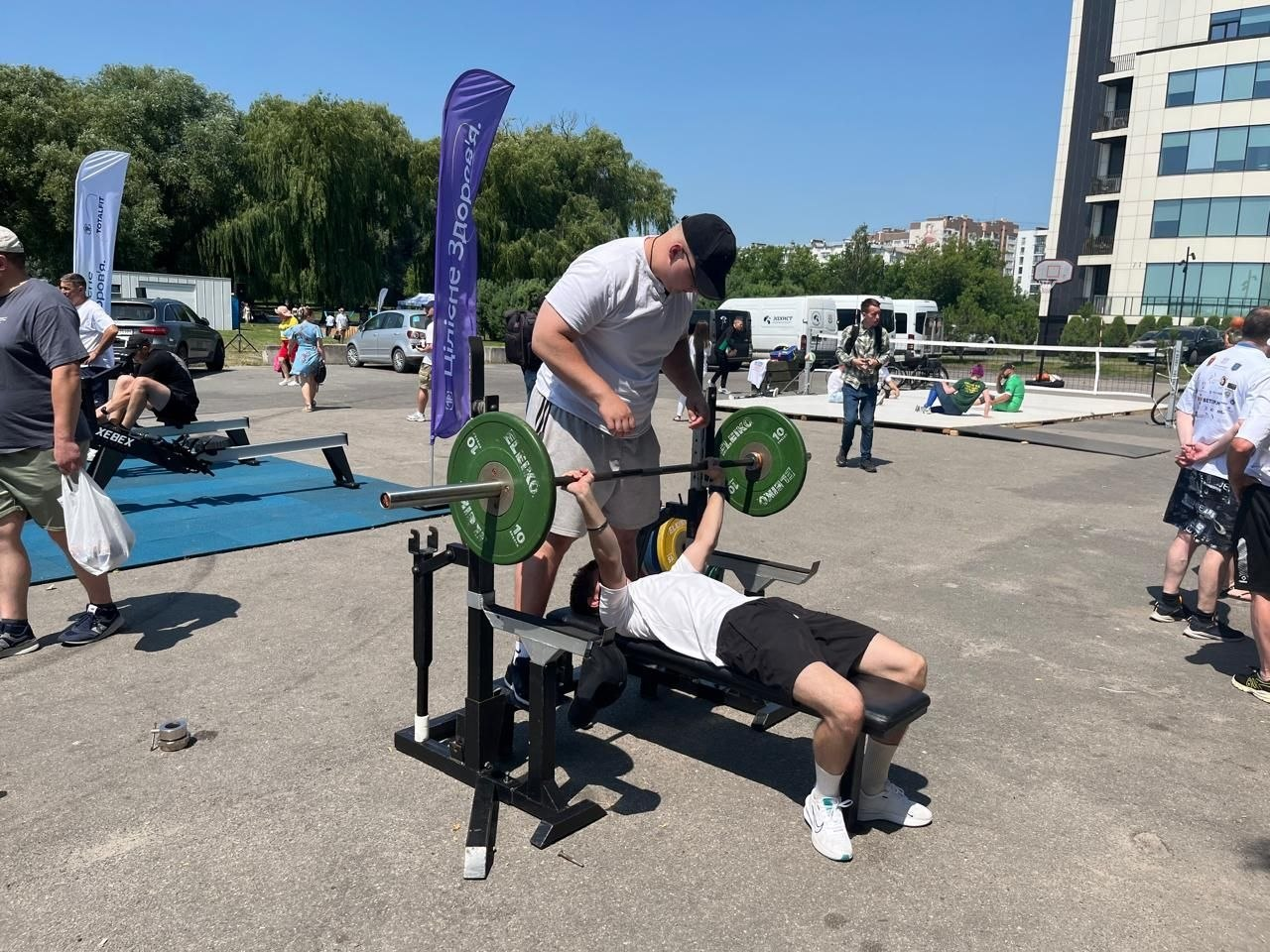 Зона силового спорту на фестивалі «Ветеран Сіті Спорт 2026» у Хмельницькому. Хлопець виконує жим штанги лежачи на лаві просто неба, а інший чоловік підстраховує його. На тлі видно тренажери для веслування, прапори з логотипами, перехожих та сучасну будівлю парку.