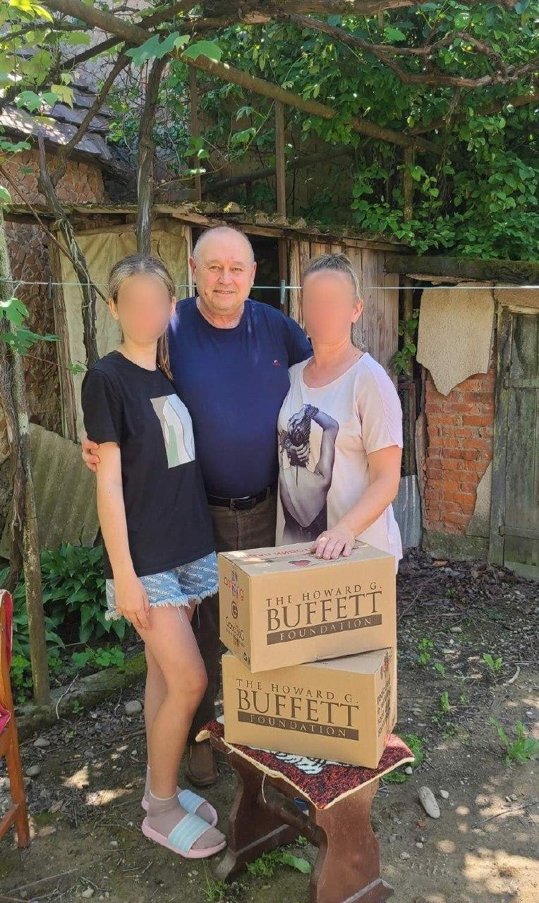 Начальник Таврійської міської військової адміністрації Микола Різак стоїть на подвір'ї поруч із двома жительками громади (їхні обличчя розмиті), які знайшли прихисток у Закарпатській області. Перед ними на дерев'яному стільці складені дві картонні коробки з гуманітарною допомогою від «The Howard G. Buffett Foundation».