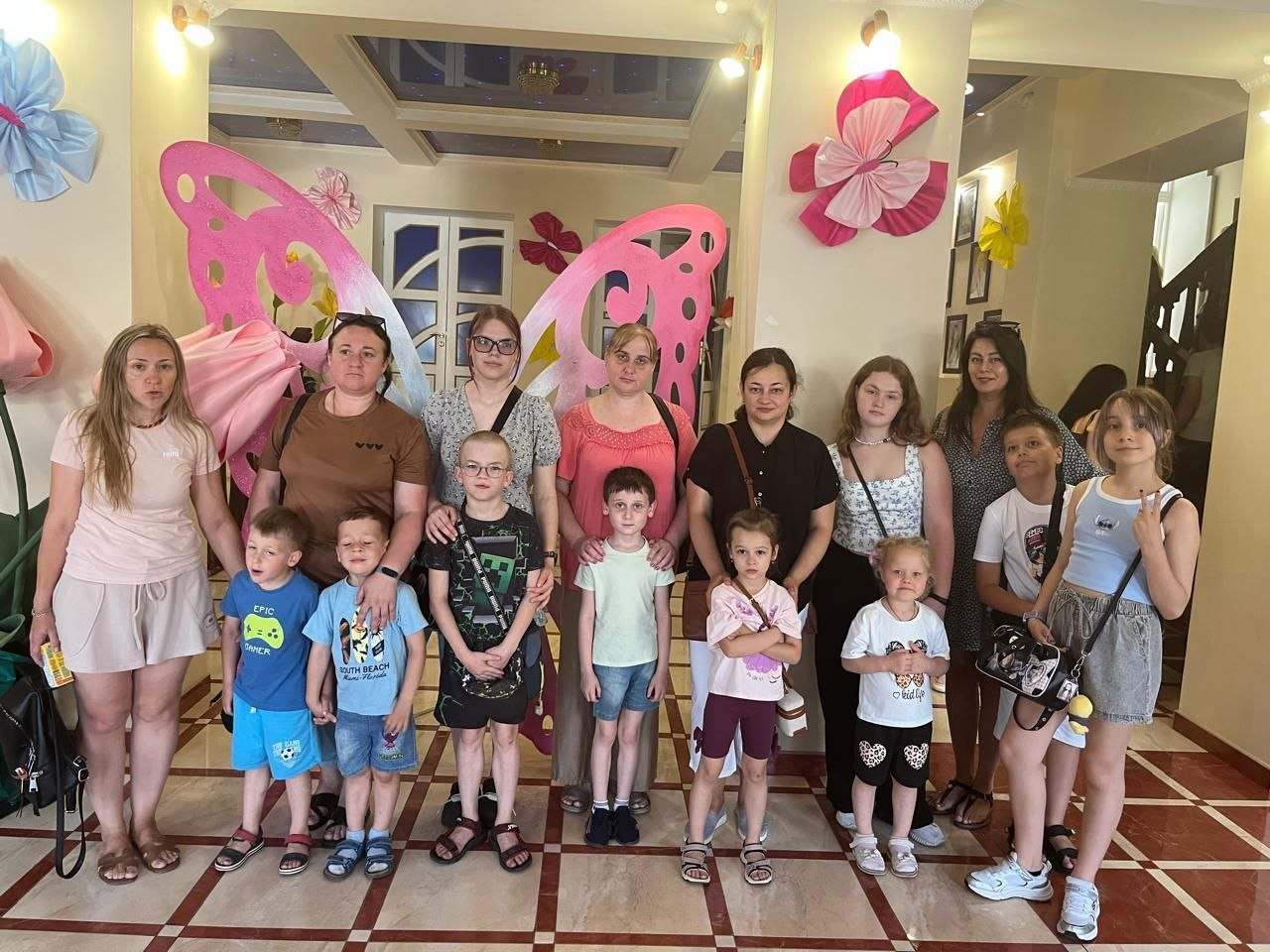 Групове фото родин-переселенців із Херсонщини у вестибюлі Хмельницького обласного академічного театру ляльок. Жінки та діти різного віку стоять у кілька рядів у літньому повсякденному одязі на тлі інтер'єру з великими декоративними рожевими метеликами та квітковими прикрасами на стінах.
