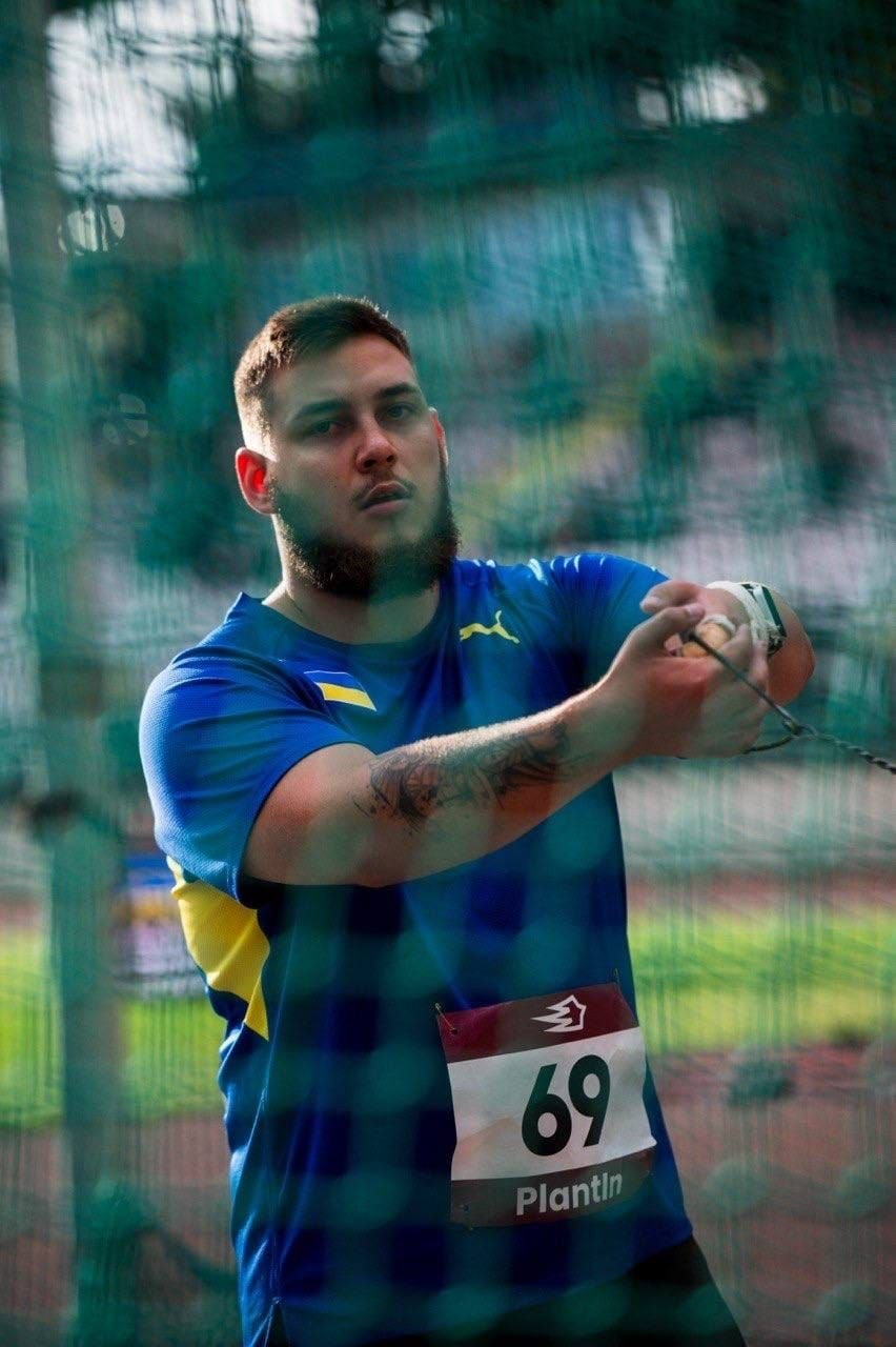 Херсонський спортсмен Микита Брагінець під час змагань із метання молота у Кропивницькому. Легкоатлет у синій футболці з українською символікою та стартовим номером 69 тримає в руках тросик молота, готуючись до кидка. Фото зроблене крізь захисну спортивну сітку.