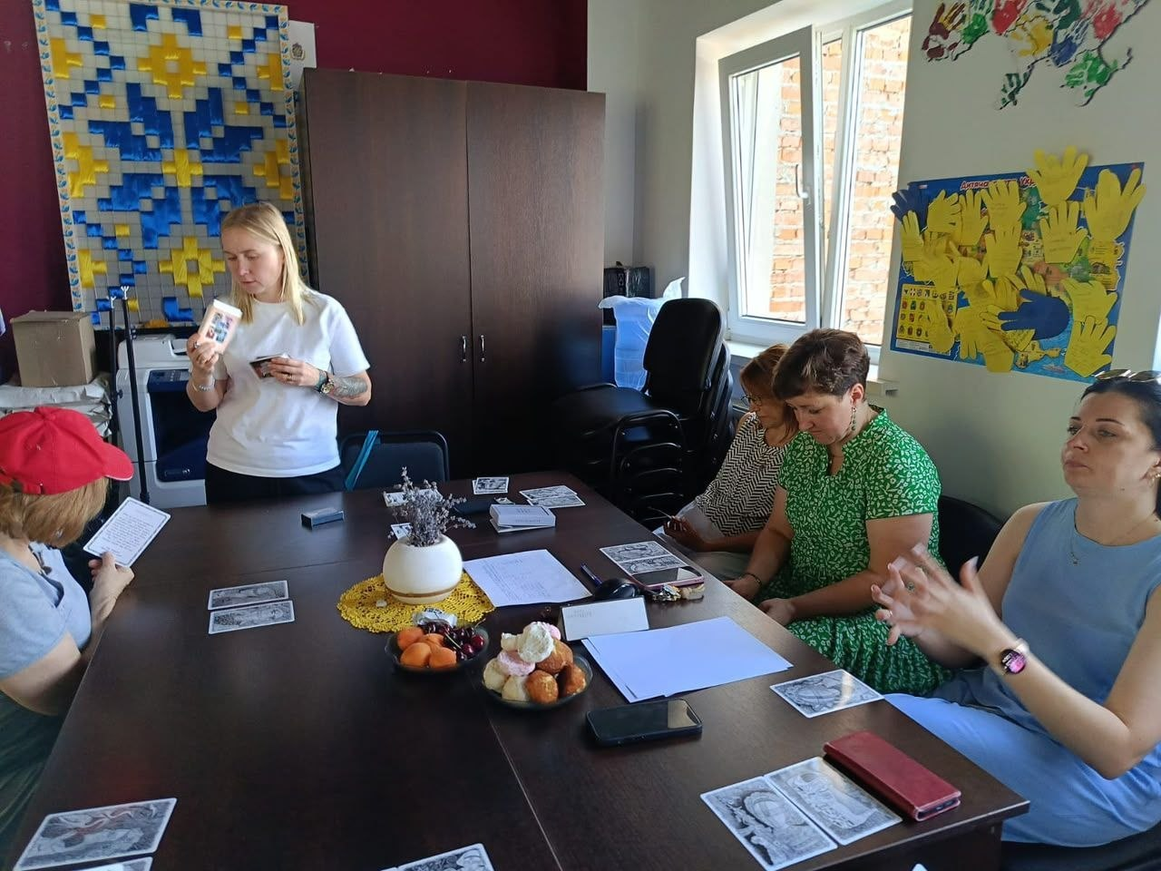 Психологічний тренінг у координаційному центрі «Вільні разом» в Івано-Франківську. Тренерка Антоніна Максимців стоїть біля столу та показує учасницям метафоричну карту. Переселенки з Херсонщини сидять навколо столу, розглядають розкладені картки із серії «Включись» та виконують практичні завдання.