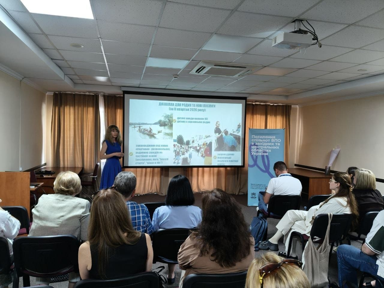 Алєся Алексєєва презентує звіт координаційного центру «Вільні разом» на регіональному заході від ГО «КримSOS». Вона виступає перед аудиторією в конференц-залі поруч із великим екраном, де відкрито слайд «Дозвілля для родин та нові послуги». Учасники сидять на стільцях і слухають спікерку, а праворуч стоїть блакитний банер заходу.
