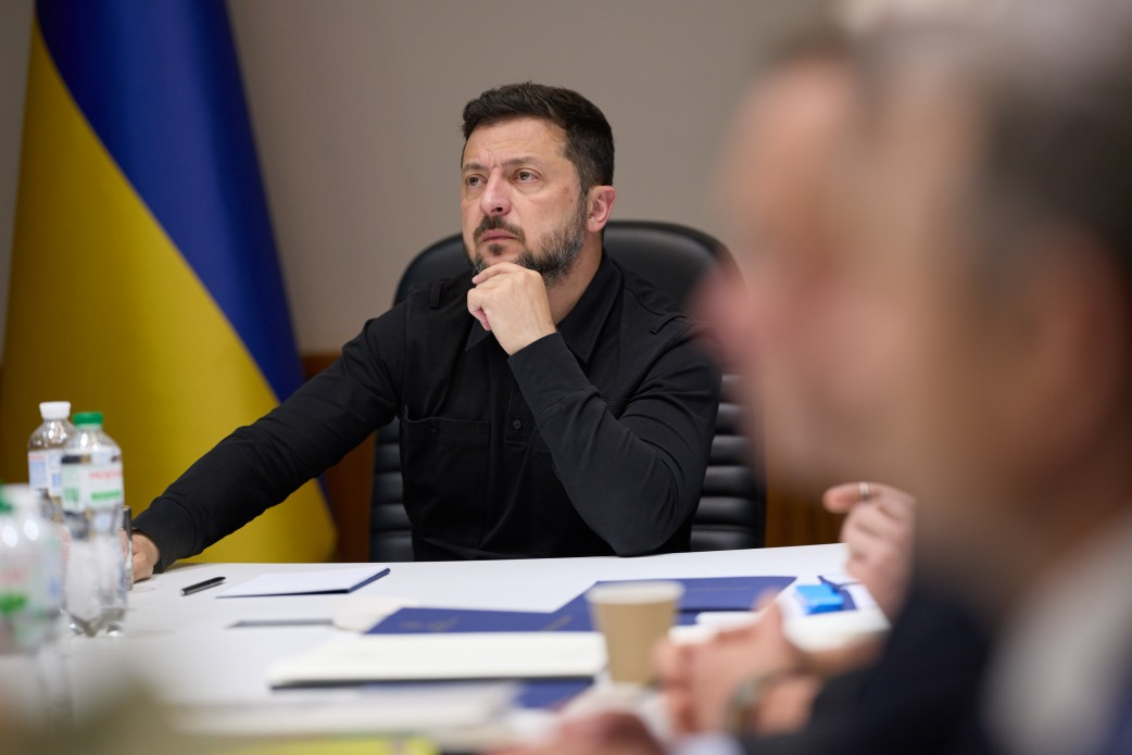 Президент України Володимир Зеленський під час спеціального засідання Ставки Верховного Головнокомандувача. Він сидить за робочим столом, підперши підборіддя рукою та зосереджено слухаючи доповідачів. На задньому плані видно Державний Прапор України, а на передньому плані — розмиті обличчя учасників зустрічі.