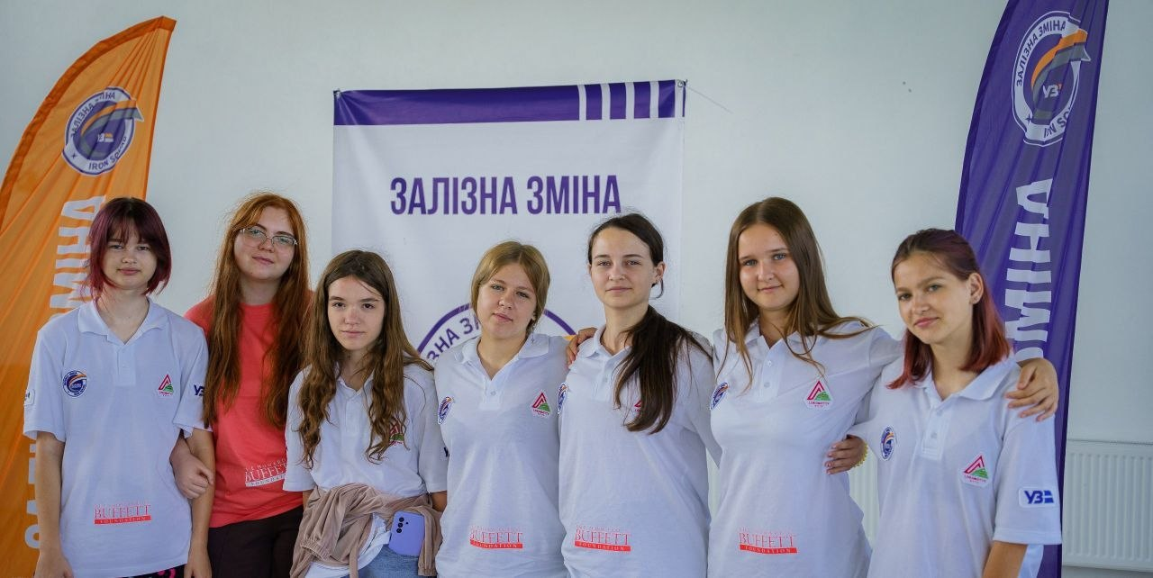 Група дівчат-учасниць проєкту «Залізна зміна» обіймаються та посміхаються на тлі фірмового банера.