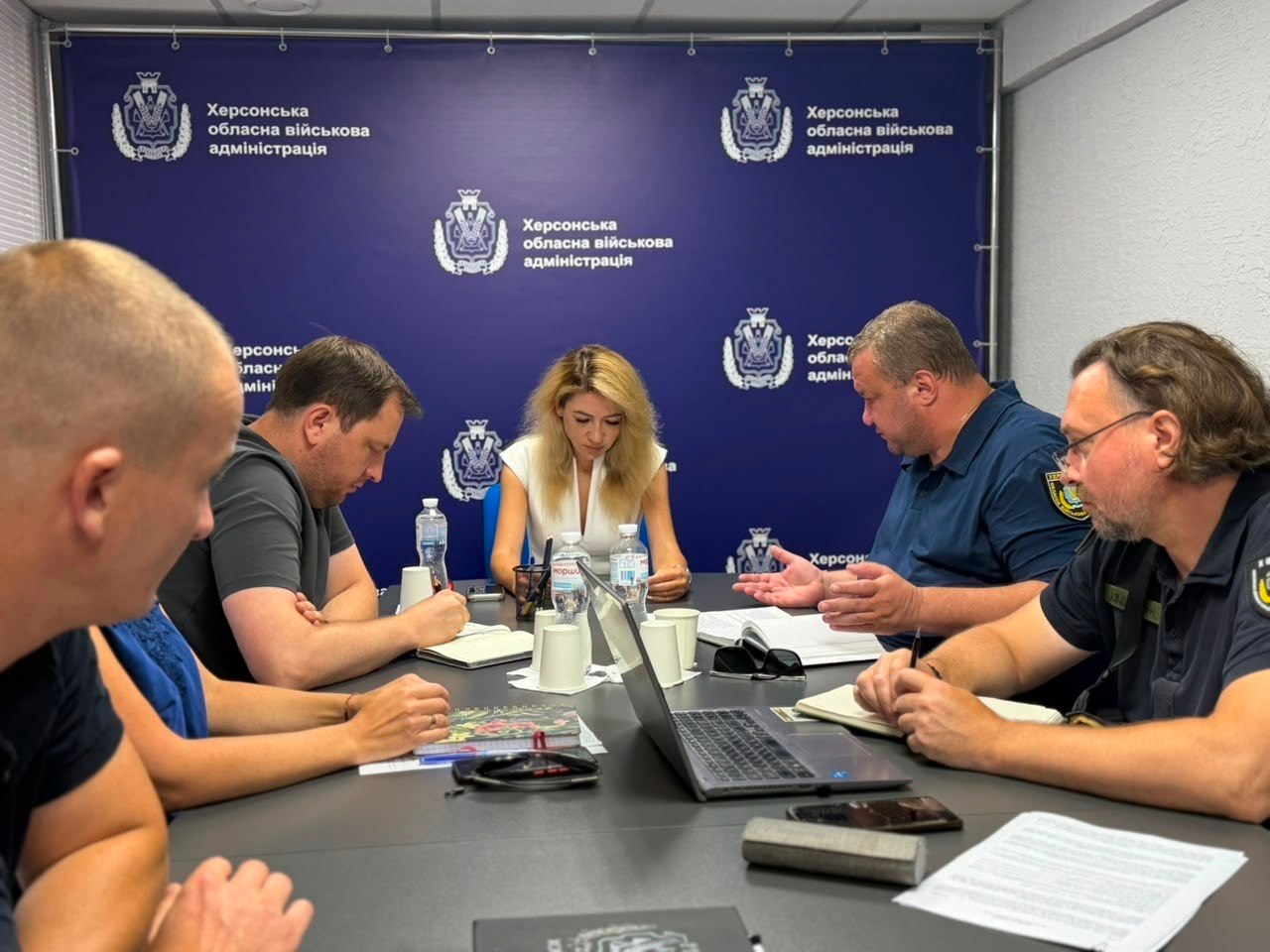 Робоча зустріч у конференц-залі Херсонської ОВА. По центру столу сидить заступниця начальника ОВА Ольга Малярчук. Ліворуч від неї, на передньому плані, сидить директор Департаменту з питань цивільного захисту та оборонної роботи ХОДА Євген Пісоцький, який робить записи у блокноті. Навпроти них сидять інші учасники наради, обговорюючи робочі документи. На задньому плані розташований синій брендований банер установи.