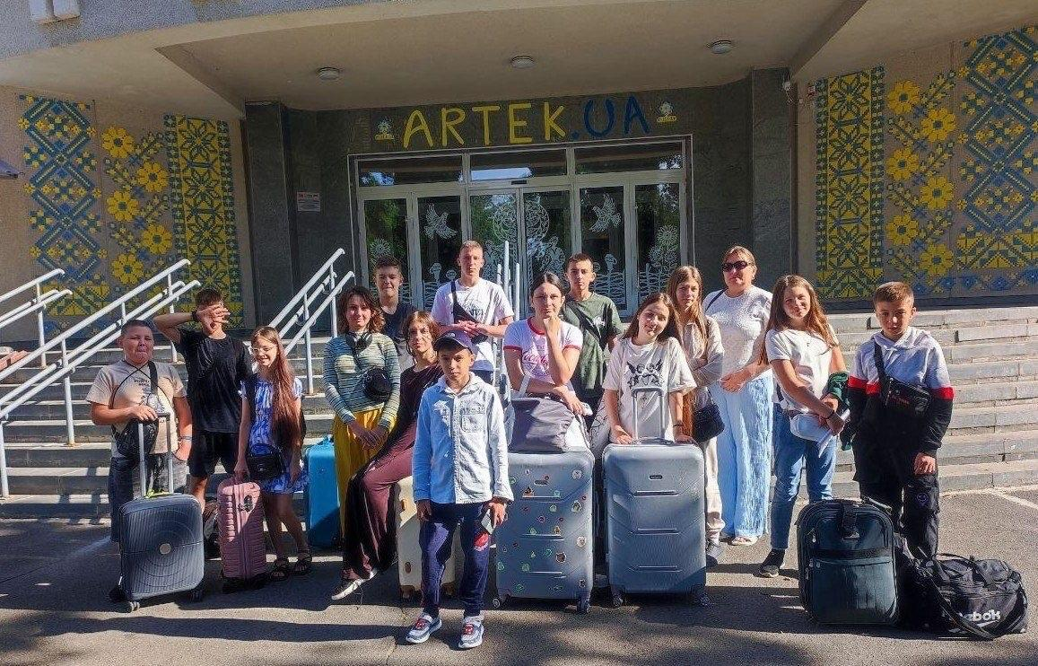 Група з дітей та підлітків разом із жінкою-супроводжуючою стоять із дорожніми валізами на вулиці біля входу до МДЦ «Артек». Учасники сьомої тематичної зміни з різних громад Херсонщини посміхаються та дивляться в кадр. Стіни будівлі навколо входу прикрашені мозаїчними панно із жовто-блакитним українським орнаментом та соняшниками.