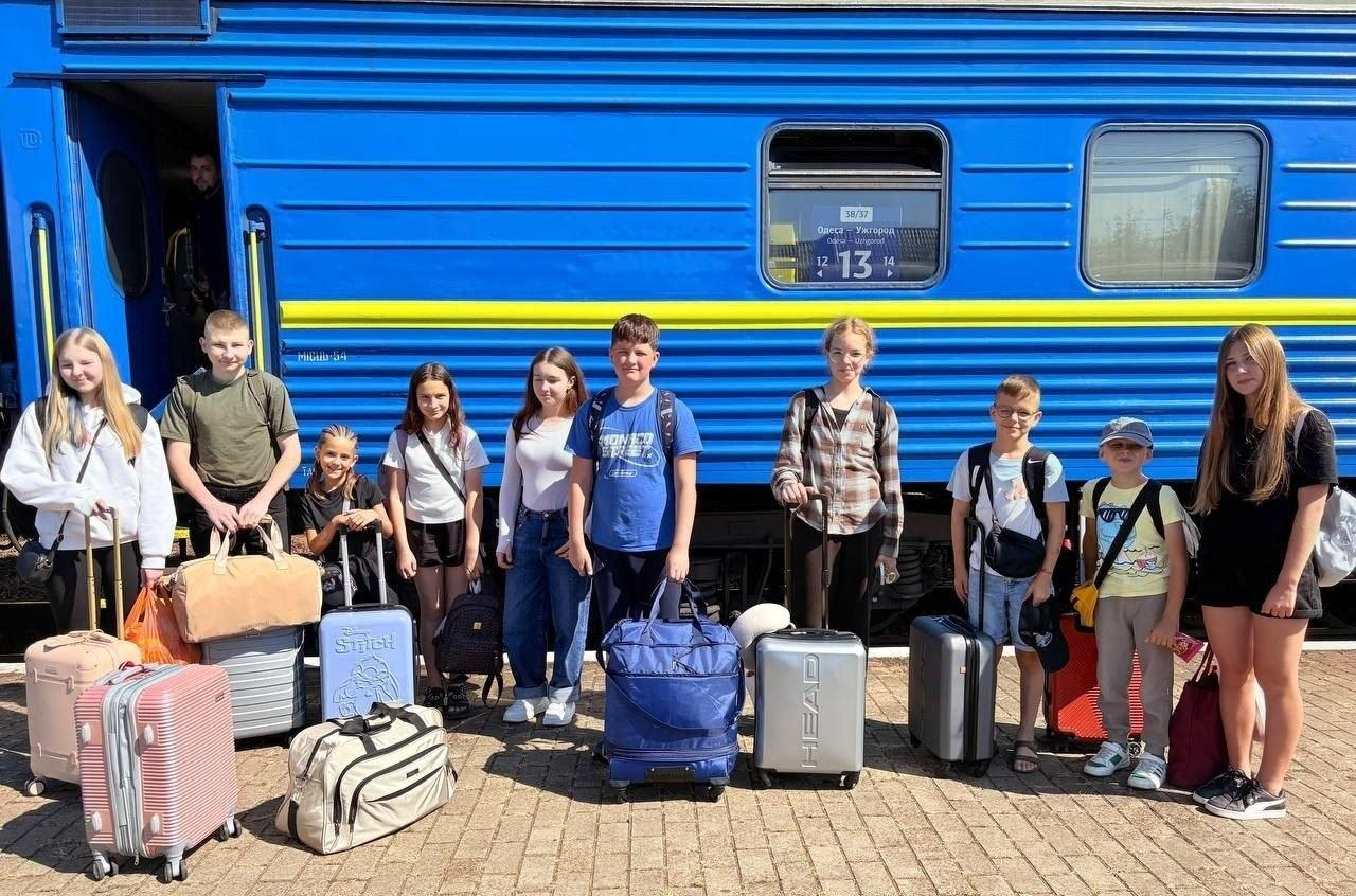 Група українських дітей та підлітків стоять із дорожніми сумками та валізами на брукованій платформі вокзалу. Вони позиють на тлі синього пасажирського вагона Укрзалізниці із жовтою смугою та табличкою маршруту «Одеса — Ужгород». Діти одягнені в літній повсякденний одяг, готові до поїздки на відпочинок.