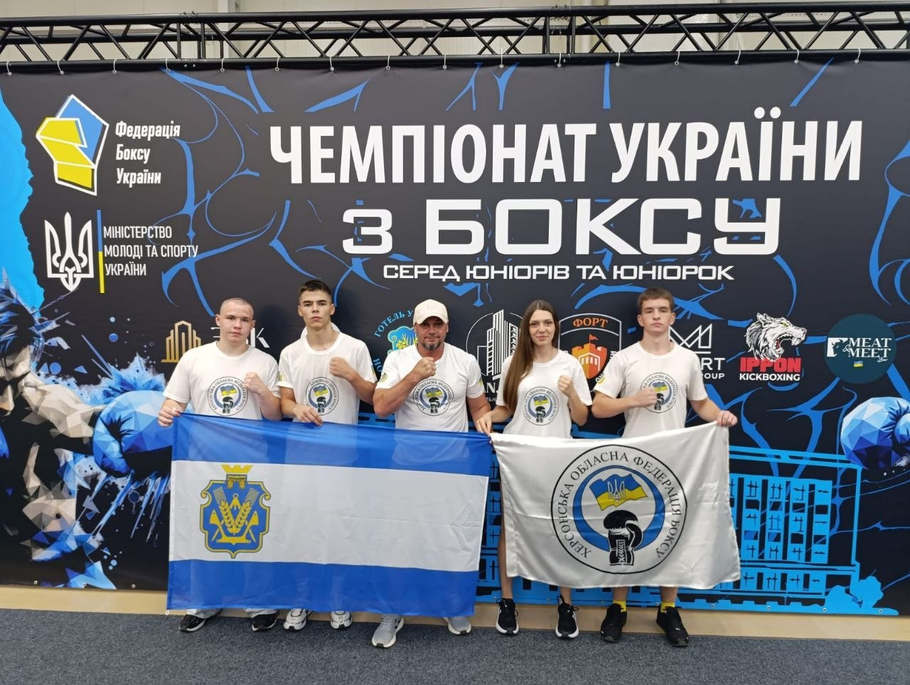 Представники Херсонської обласної федерації боксу стоять разом на тлі офіційного банера «Чемпіонат України з боксу серед юніорів та юніорок» в Ужгороді, тримаючи прапори своєї організації та регіону.