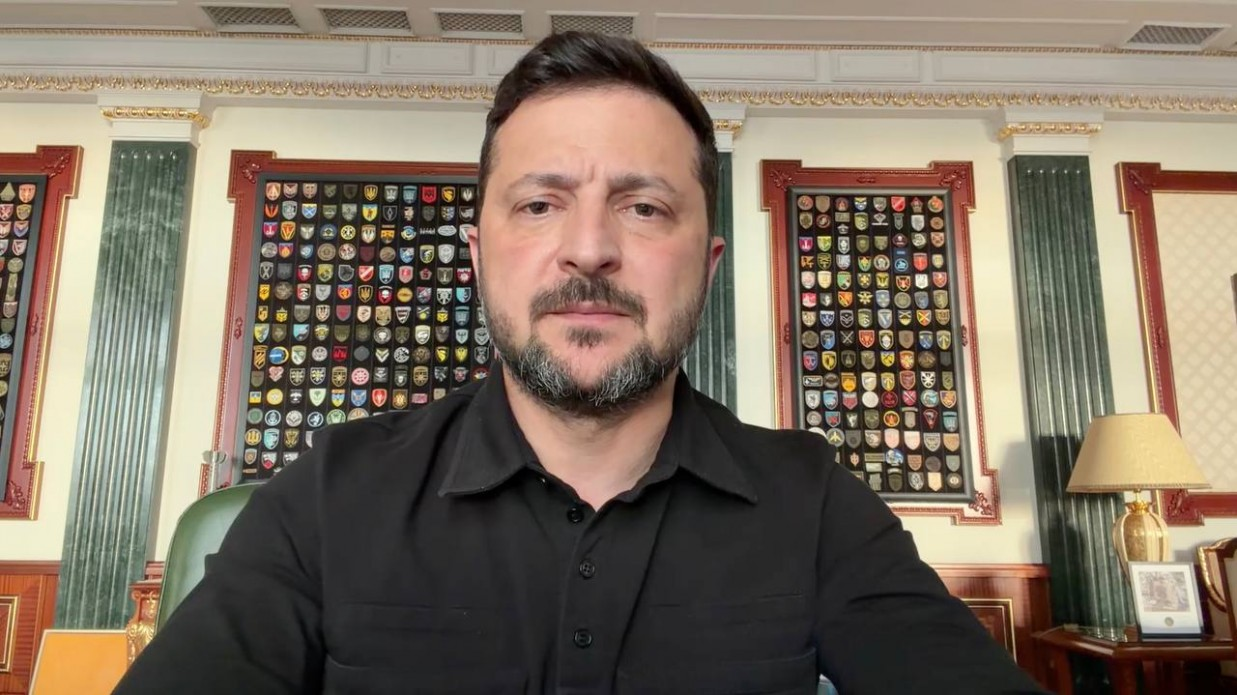 Президент України Володимир Зеленський у чорній сорочці під час звернення на тлі стіни з шевронами українських військових підрозділів.