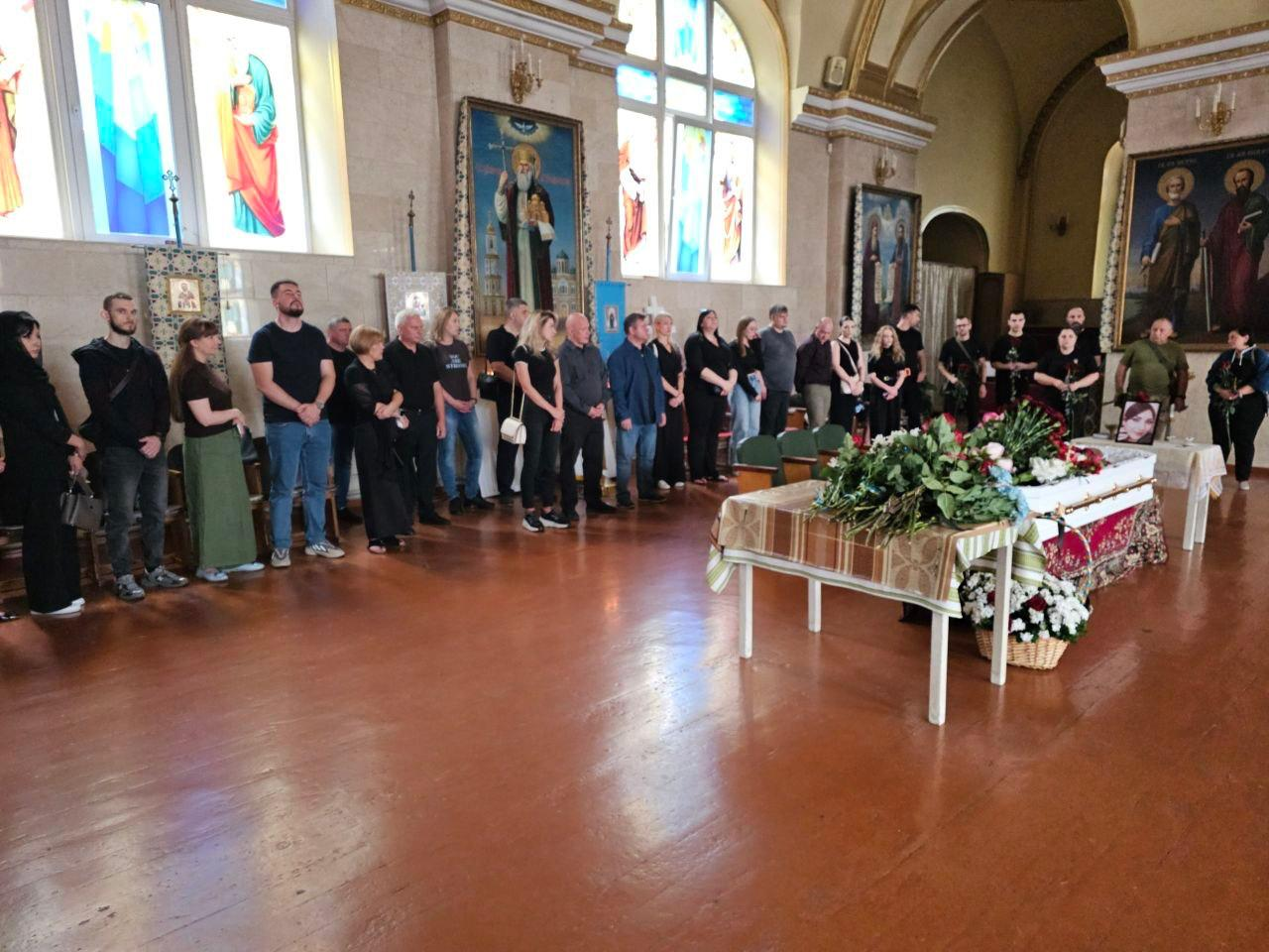 Церемонія прощання з працівницею Херсонської обласної військової адміністрації Анжелікою Пєрєвєрзєвою у храмі. Люди у чорному вбранні стоять уздовж стіни, а в центрі зали розташована вкрита квітами труна. На задньому плані видно церковні ікони та вітражні вікна.