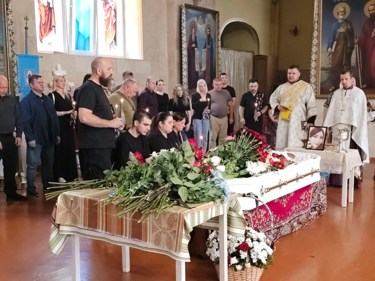 Церемонія відспівування у храмі працівниці Херсонської ОВА Анжеліки Пєрєвєрзєвої. Священнослужителі проводять службу біля вкритої квітами труни, навколо якої зібралися родичі та колеги із запаленими свічками в руках.
