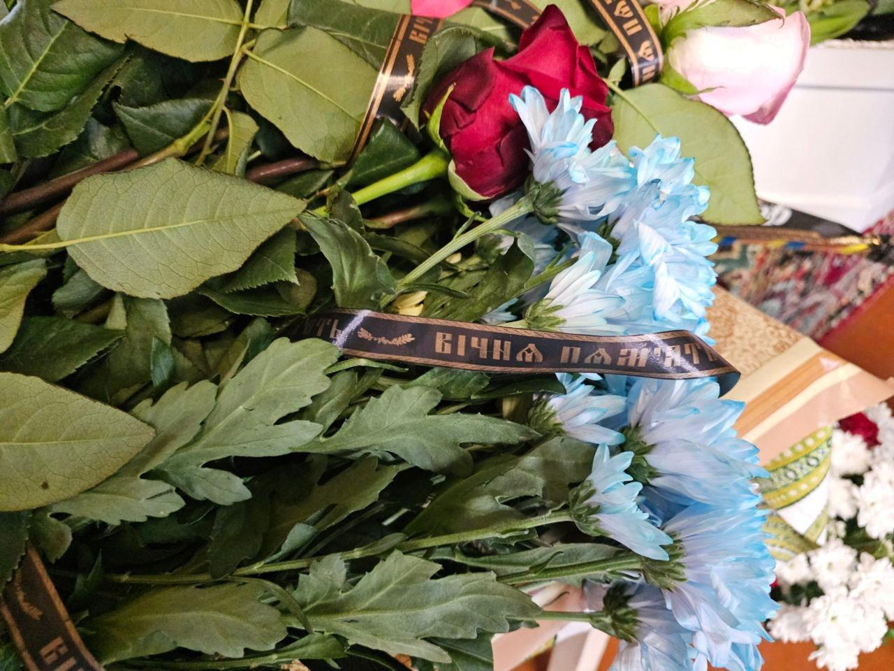 Великий букет жалобних квітів, серед яких блакитні хризантеми та червоні троянди, перев'язаний чорною стрічкою з написом «Вічна пам'ять».
